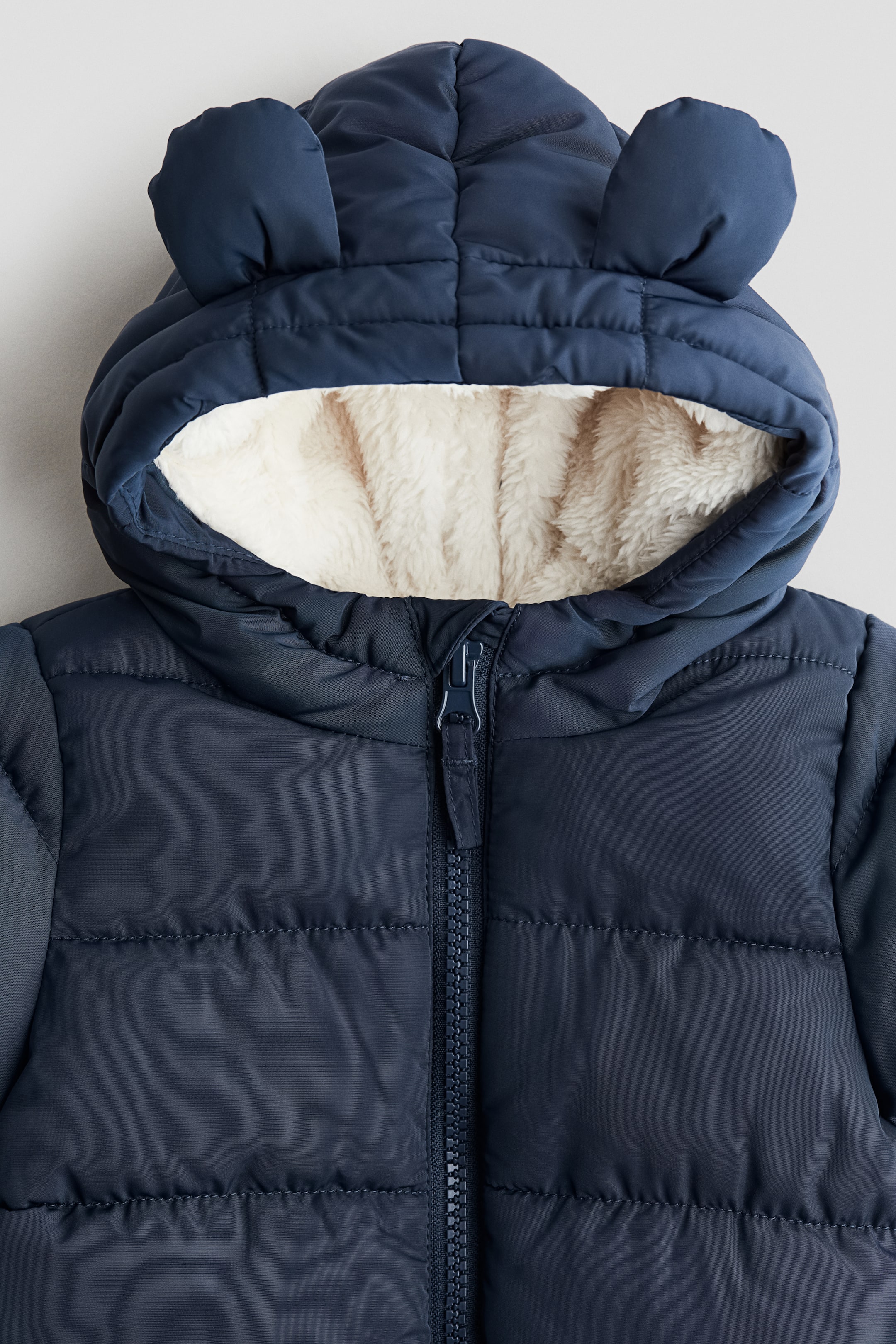 View larger image: A navy blue, pile-lined snowsuit is presented from the FRONT, featuring a padded design with horizontal quilting. Its attached hood is lined with cream-colored pile and is displaying two small, rounded ears on top, while a navy blue zipper is running down the center front.