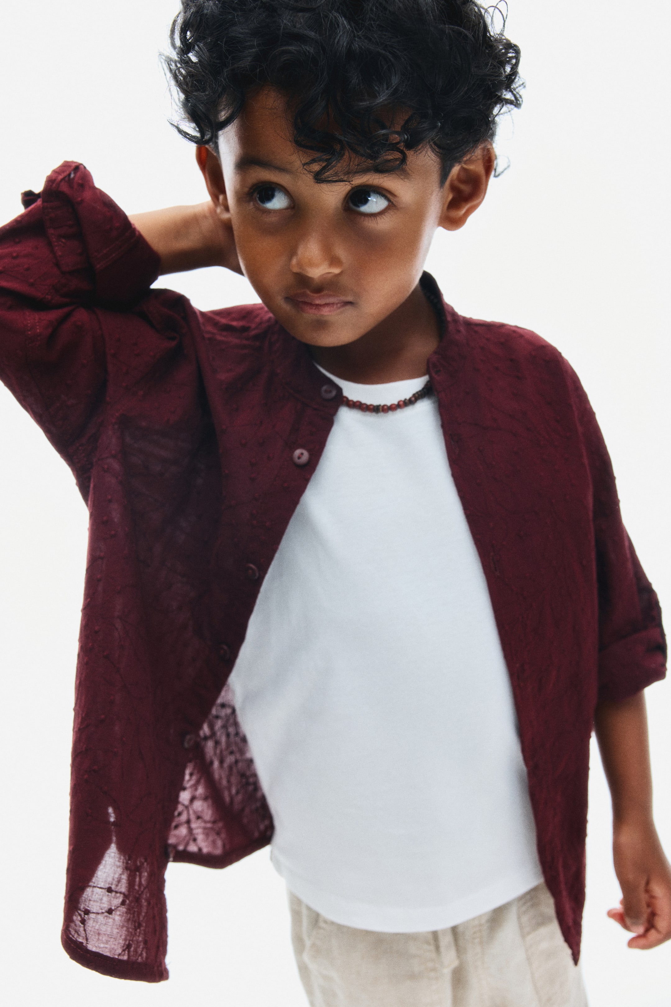 View larger image: A burgundy grandad shirt in a textured, patterned cotton weave is shown from the front, featuring a band collar and front button fastening. The shirt is worn open over a white T-shirt, highlighting its lightweight fabric and long sleeves rolled up to the elbows.