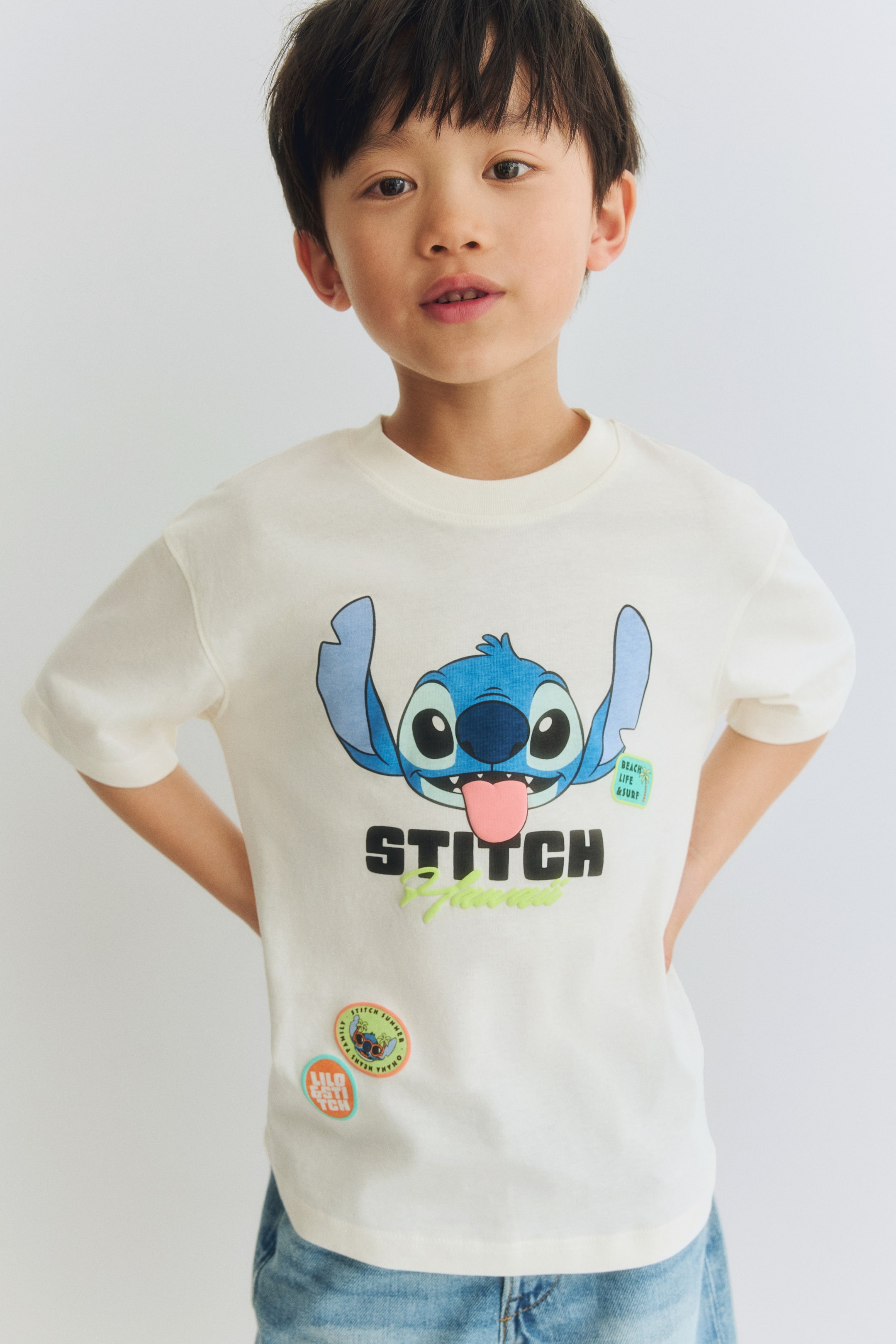 View larger image: An oversized t-shirt crafted from soft, printed cotton jersey is displayed from the front, showcasing a large graphic of Stitch accompanied by the text "STITCH" and "Hawaii," along with several small graphic patches. It features a ribbed trim around the neckline and has dropped shoulders.