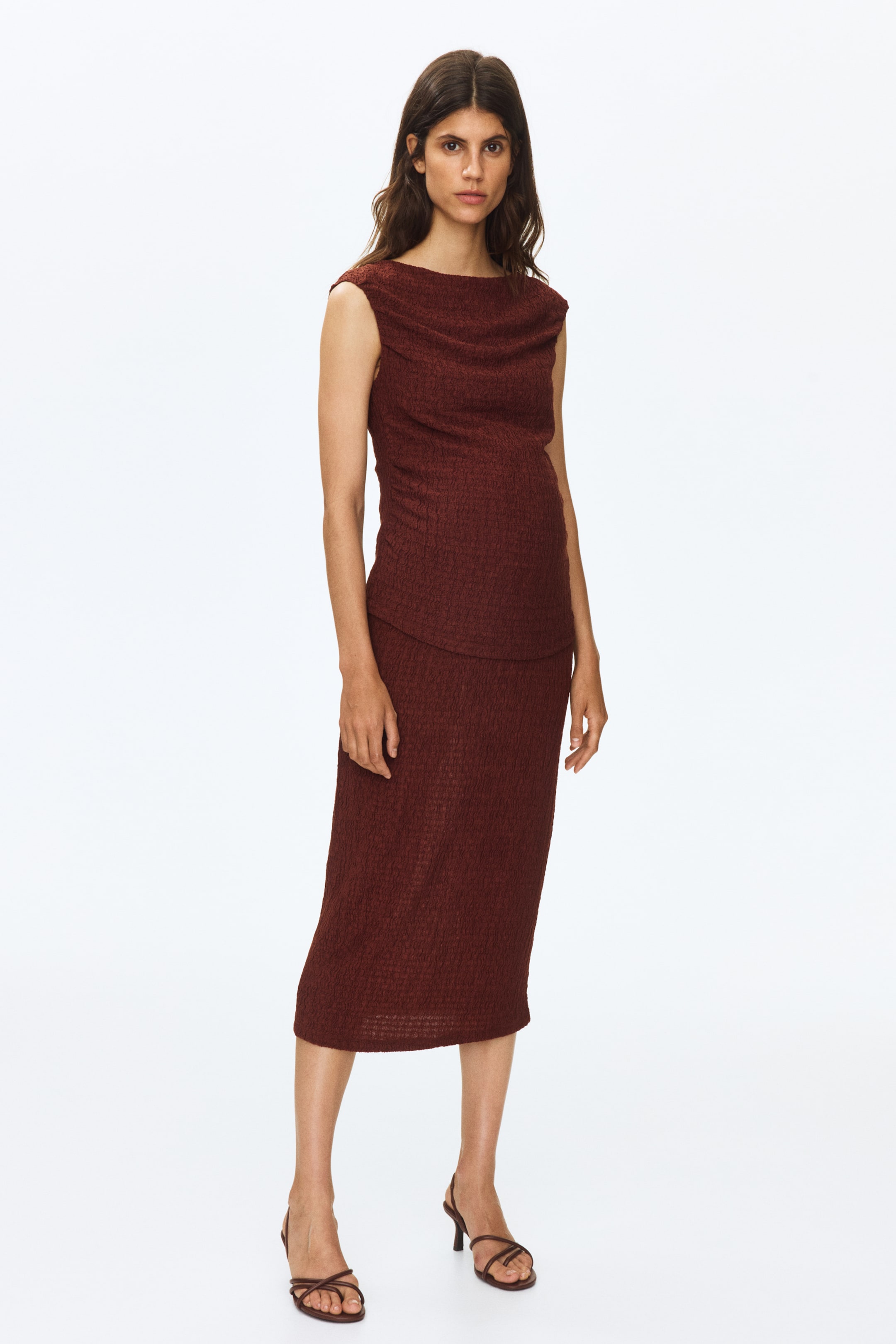 View larger image: A reddish-brown set, crafted from textured stretch jersey, is showcased by the model; the fitted, sleeveless top highlights a boat neckline with a draped effect, while the calf-length skirt features a high waist. The model complements the look with dark strappy heels.