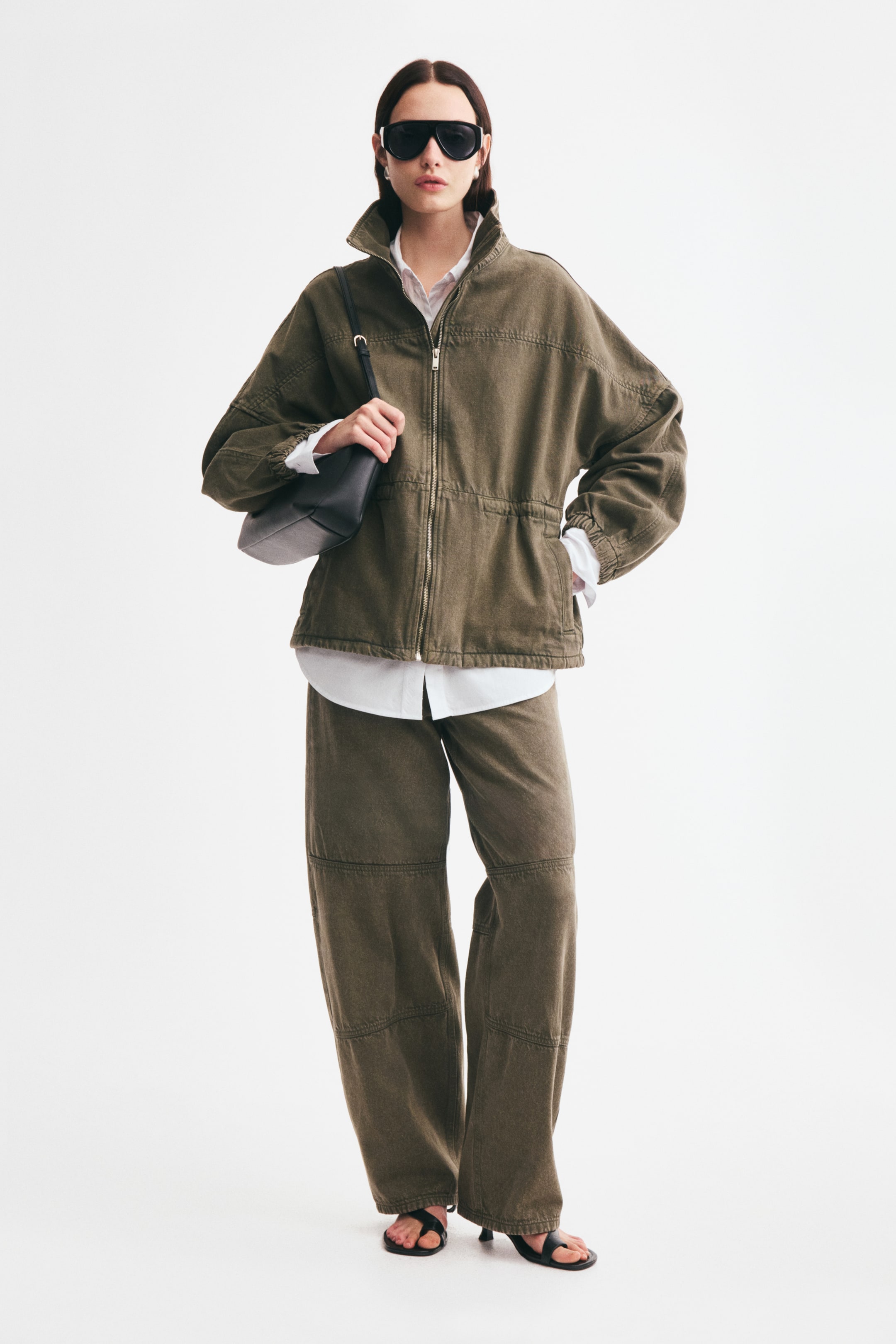 View larger image: An oversized jacket in olive-green cotton denim is worn by the model, layered over a white collared shirt and paired with matching wide-leg trousers. The jacket features a funnel collar, a front zip, dropped shoulders, and a drawstring waist, with the model holding the strap of a black shoulder bag.