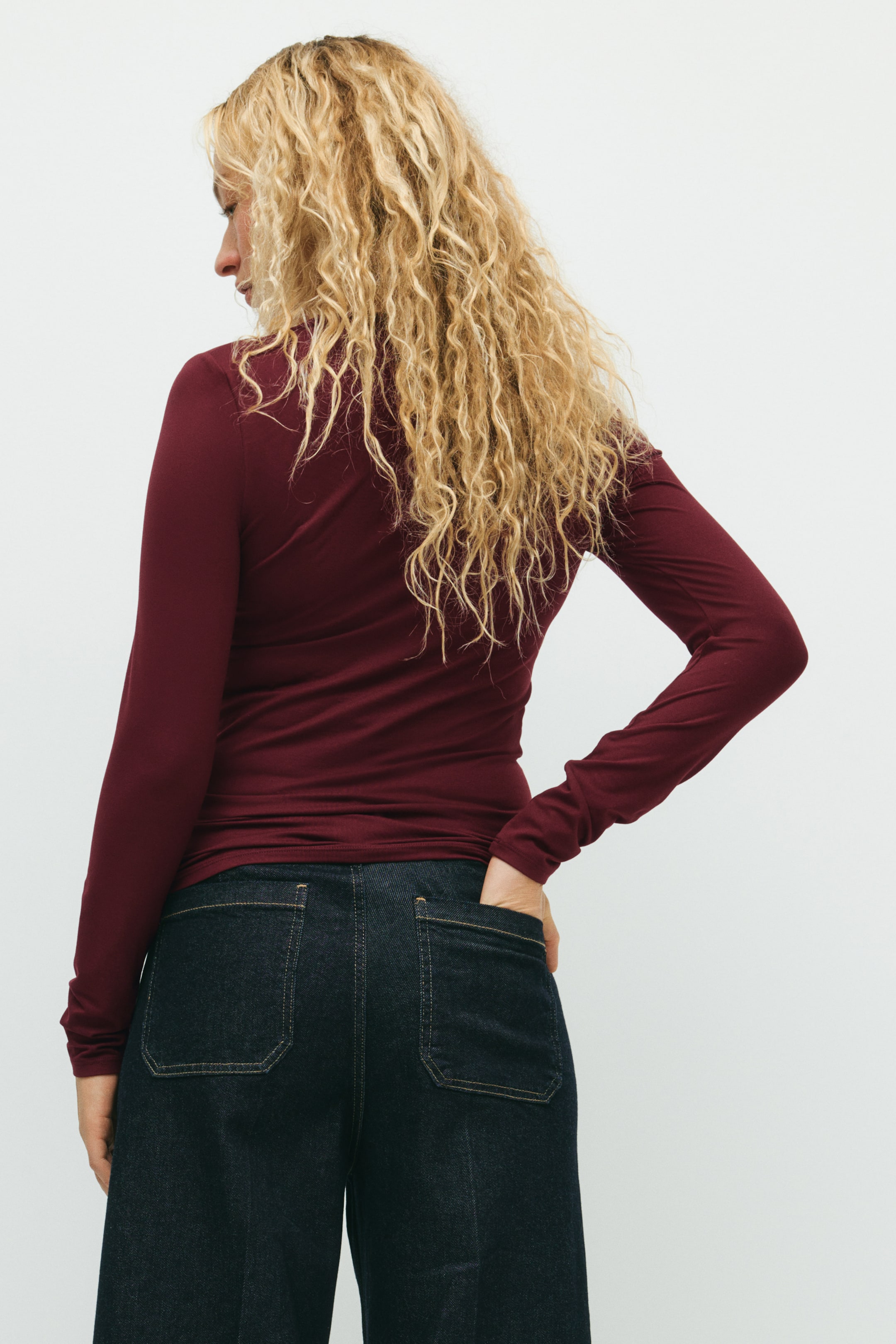 Ampliar la imagen: La modelo lleva puesto un top ajustado de jersey rojo oscuro con mangas largas, junto con pantalones oscuros y una mano descansando en el bolsillo trasero.