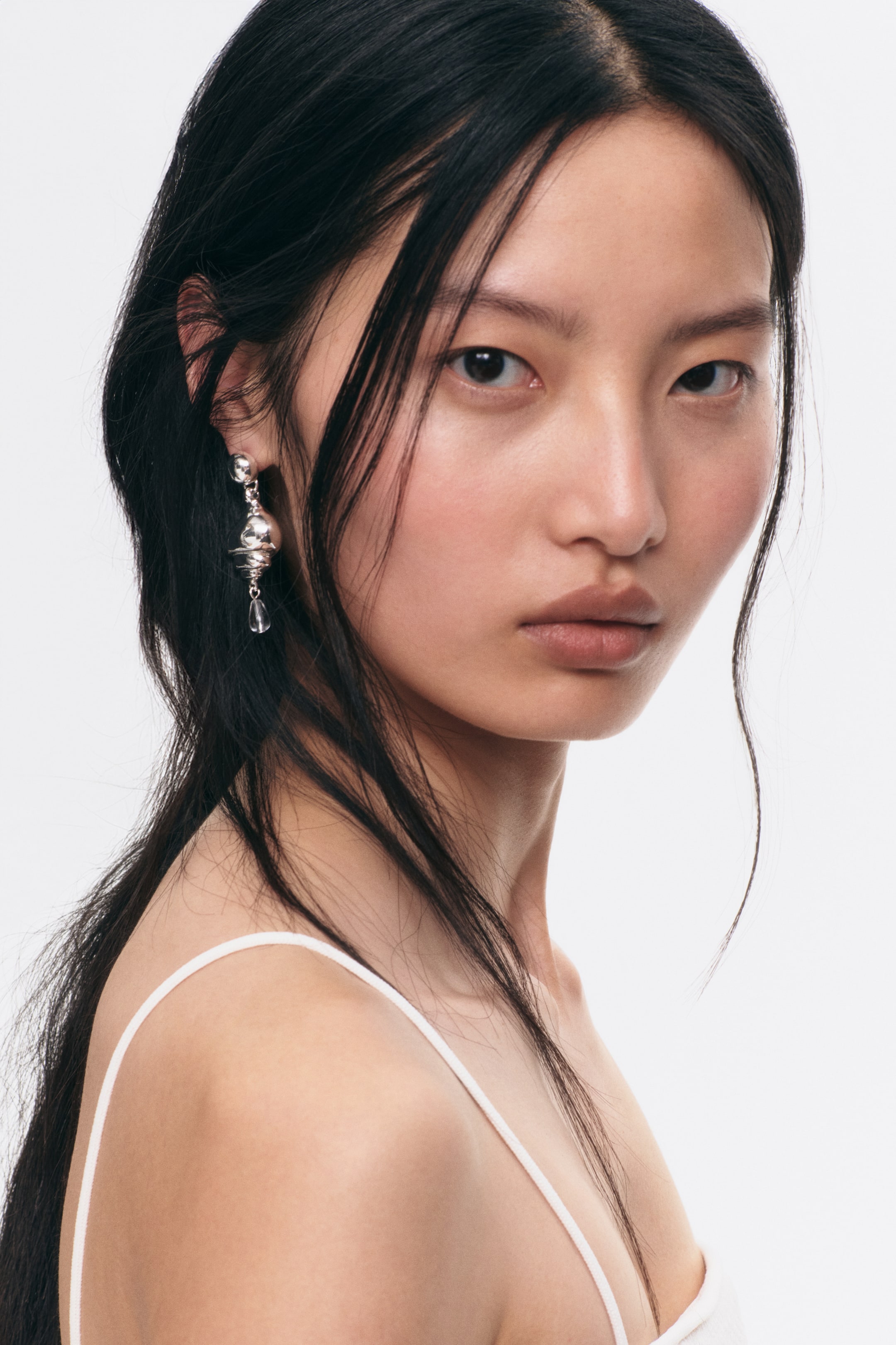 Agrandir l'image: La boucle d'oreille est un modèle pendante de ton argenté, comportant un petit clou sphérique, une perle de ton argenté de forme organique plus grande, et un pendentif en forme de goutte d'eau transparent. Elle est suspendue verticalement à une oreille.