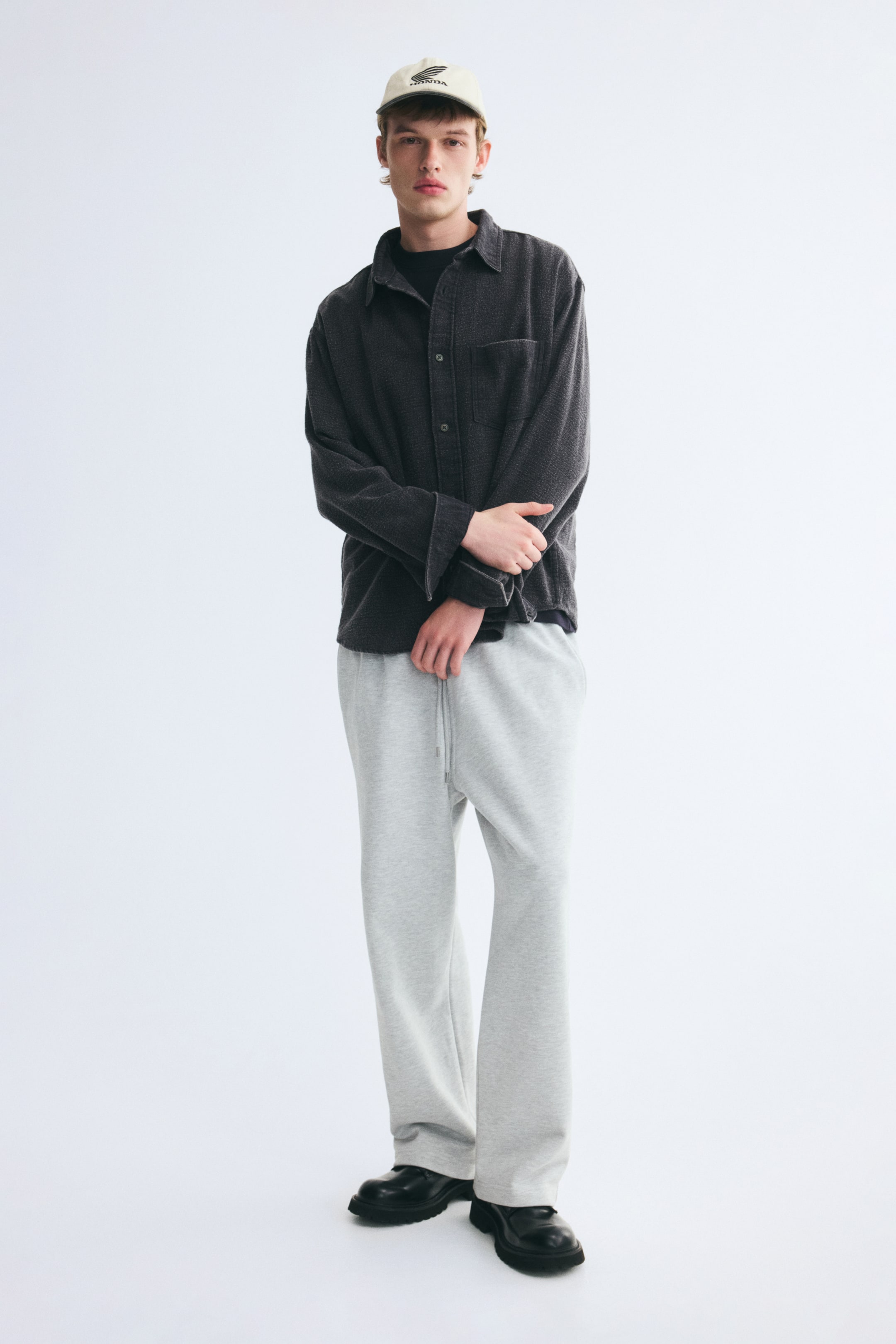 View larger image: An overshirt in dark grey cotton twill with a distressed look is worn by the model, layered over a black t-shirt and styled with light grey sweatpants. The overshirt features a collar, front button closure, a single patch chest pocket, and long sleeves with button cuffs.