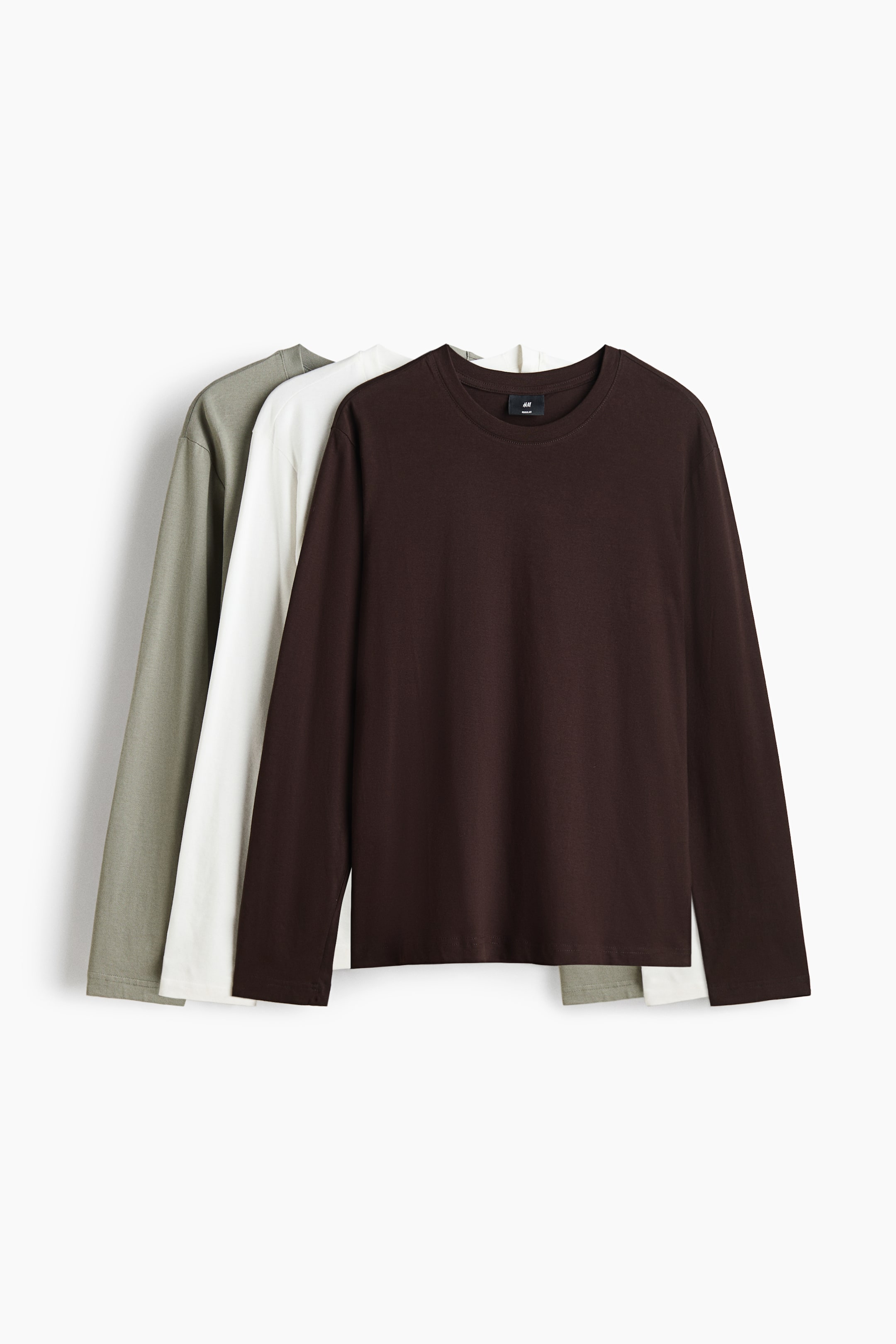 View larger image: Three long-sleeved cotton jersey tops are hanging layered from front to back in a front-facing view, showing their classic silhouette with a ribbed crew neckline, long sleeves, and a straight-cut hem in dark brown, white, and olive green.