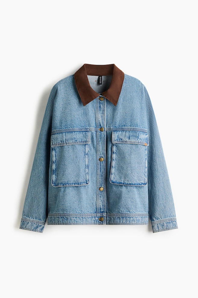 En lysvasket denimjakke vises fra forsiden, med en kontrasterende brun krave og to store påsyede lommer på brystet. Den er designet med en knaplukning foran ved hjælp af messingfarvede knapper. - 1