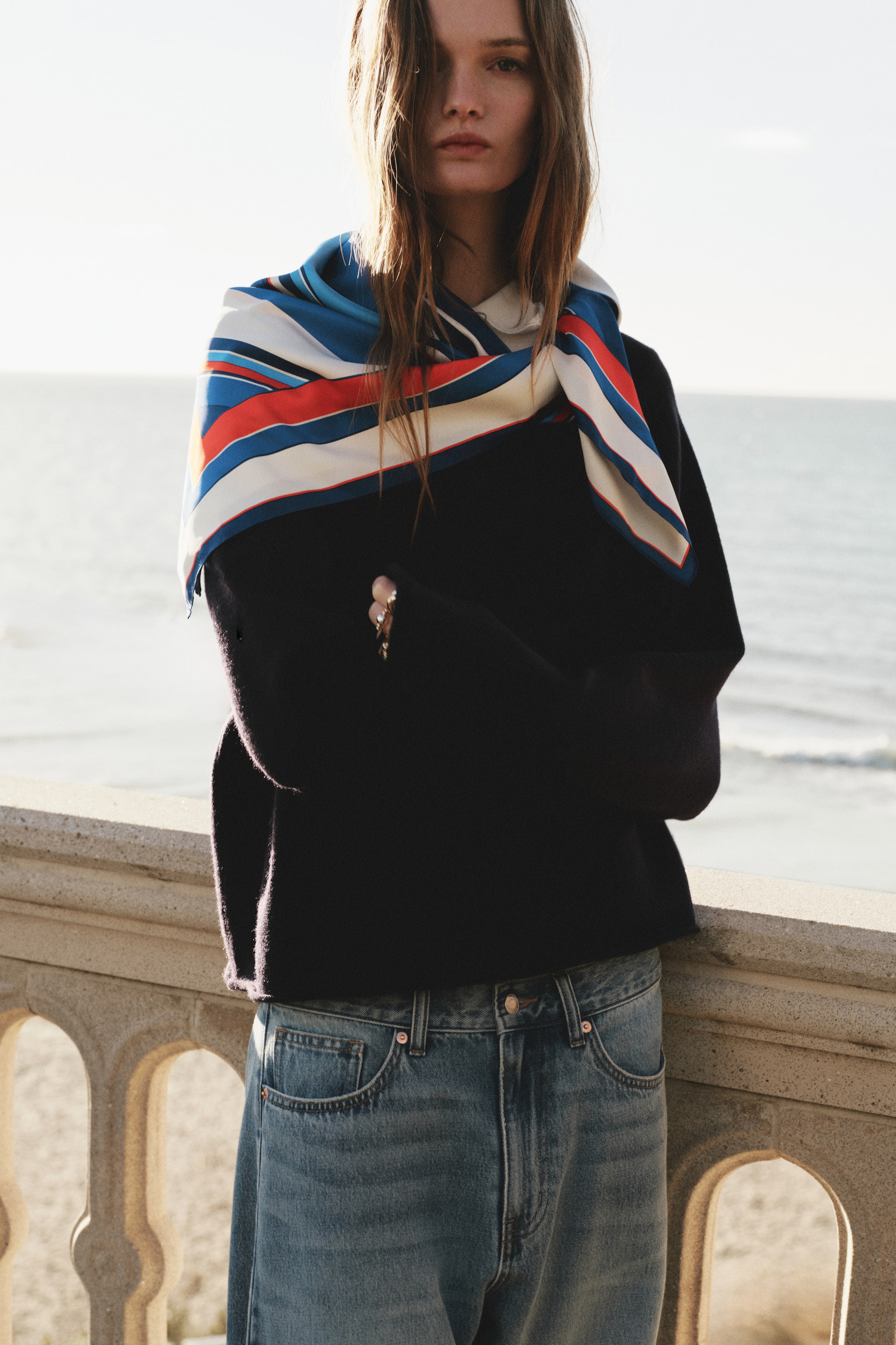 View larger image: A multicolored striped scarf in blue, white, red, and orange is draped over a dark navy, long-sleeve sweater. The sweater is paired with light-wash denim jeans that feature visible front pockets and belt loops.