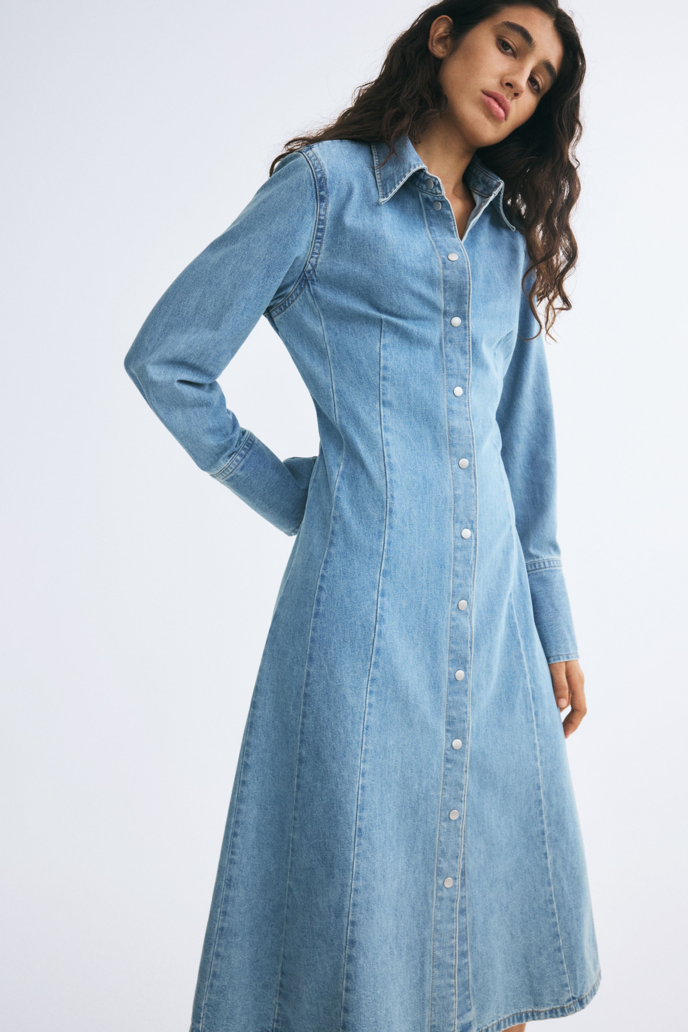 A light-wash denim midi dress is shown from the front, featuring a classic point collar, full-length snap-button closure, and long sleeves with wide, structured cuffs. Vertical seams run down the front of the dress, defining its structured, tailored silhouette.