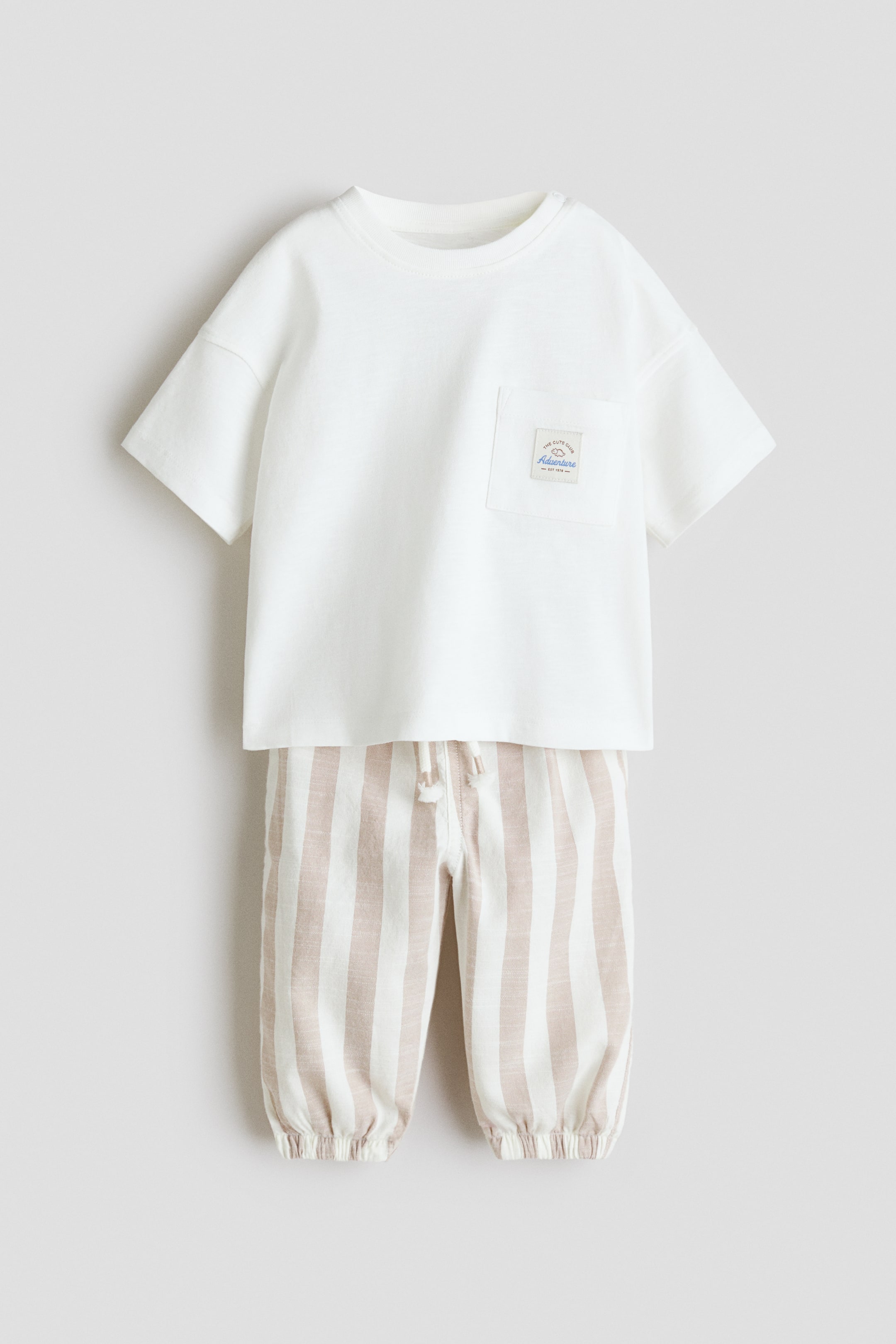 Agrandir l'image: L'ensemble 2 pièces en mélange de lin comprend un t-shirt crème à manches courtes avec un col rond et une poche décorative carrée sur la poitrine gauche. Le pantalon assorti présente des rayures verticales crème et beige, une taille élastique avec cordon de serrage, et des poignets froncés aux chevilles.