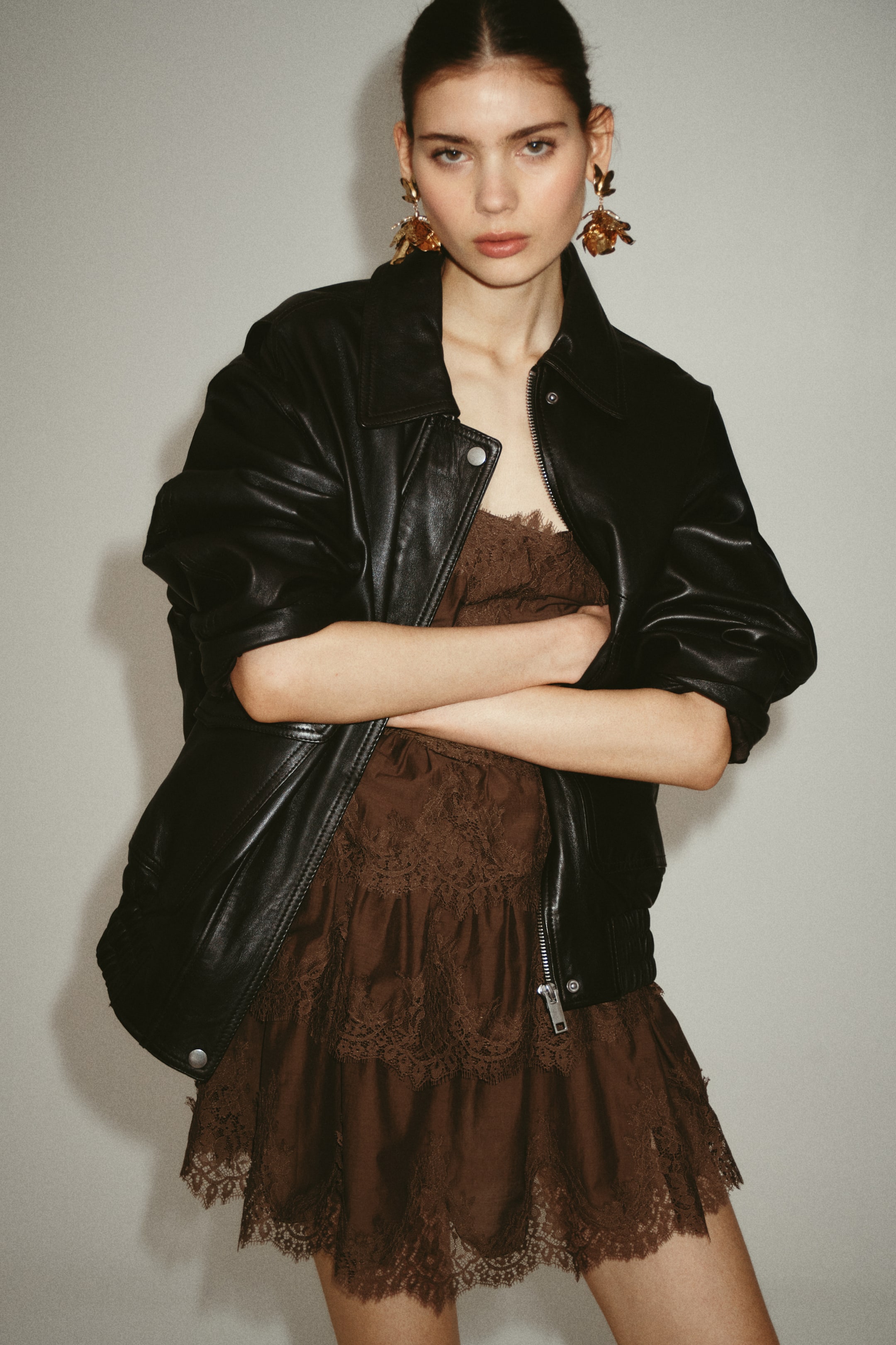 View larger image: The model is wearing a short brown woven dress with flounced, lace-trimmed tiers and a straight neckline. The dress is layered under an open black leather jacket, and the model has her arms crossed.