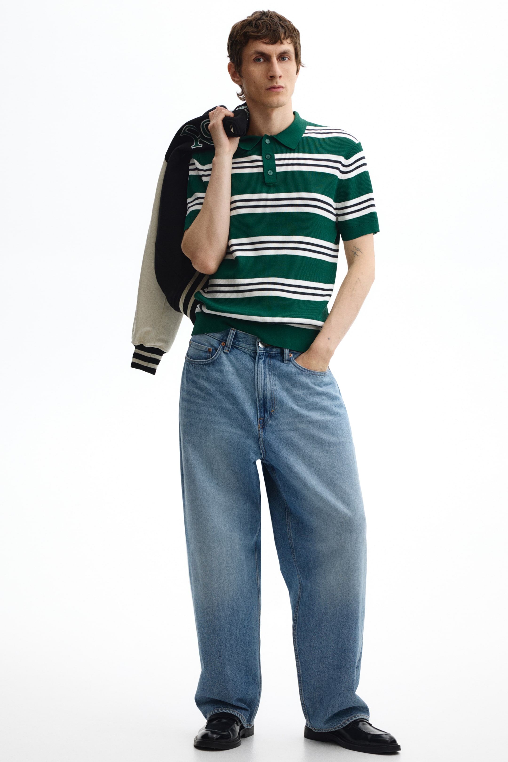 Visa större bild: En grön- och vit horisontellt randig, stickad pikétröja med spetsig krage, knappslå med tre knappar och korta ribbade ärmar bärs tillsammans med ljusblå vida jeans och svarta polerade skor. En krämfärgad och svart varsityjacka med randig ribbad mudd hänger över vänster axel.