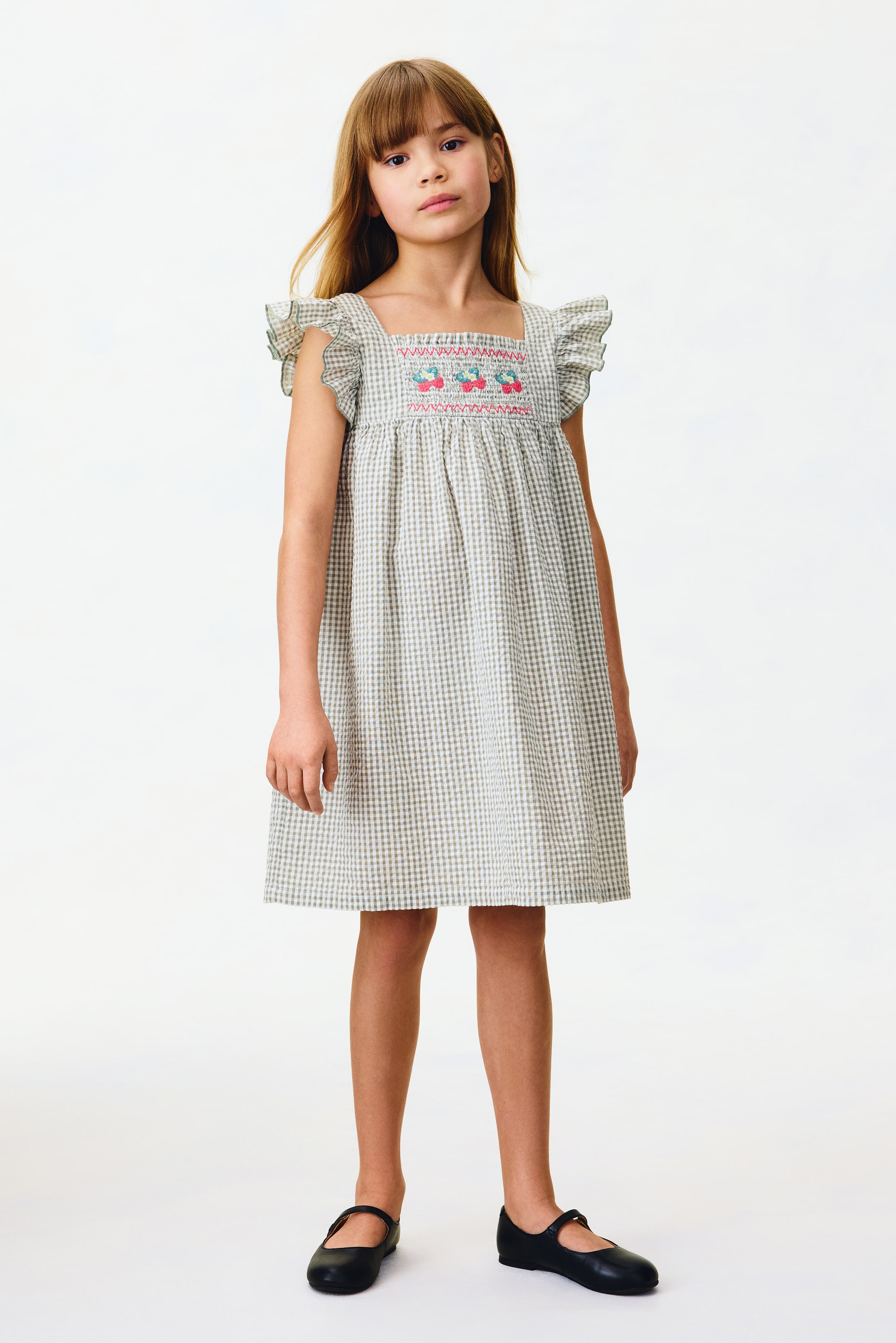 Größeres Bild anzeigen: Ein Kleid wird von vorne präsentiert, mit einem hellgrünen und weißen Vichy-Karo-Muster, einem quadratischen Ausschnitt und Flatterärmeln. Es zeigt ein gesmoktes Oberteil mit bunter Blumenstickerei und Raffungen oberhalb der Taille, die in einen ausgestellten Rock übergehen.
