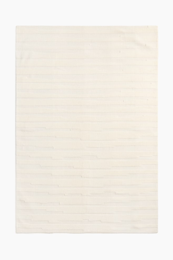 A large, woven rug showcases a cream colour and a textured pattern. This pattern consists of horizontal woven stripes, with some sections appearing raised and others flat, creating a subtle relief across its front surface. - 1