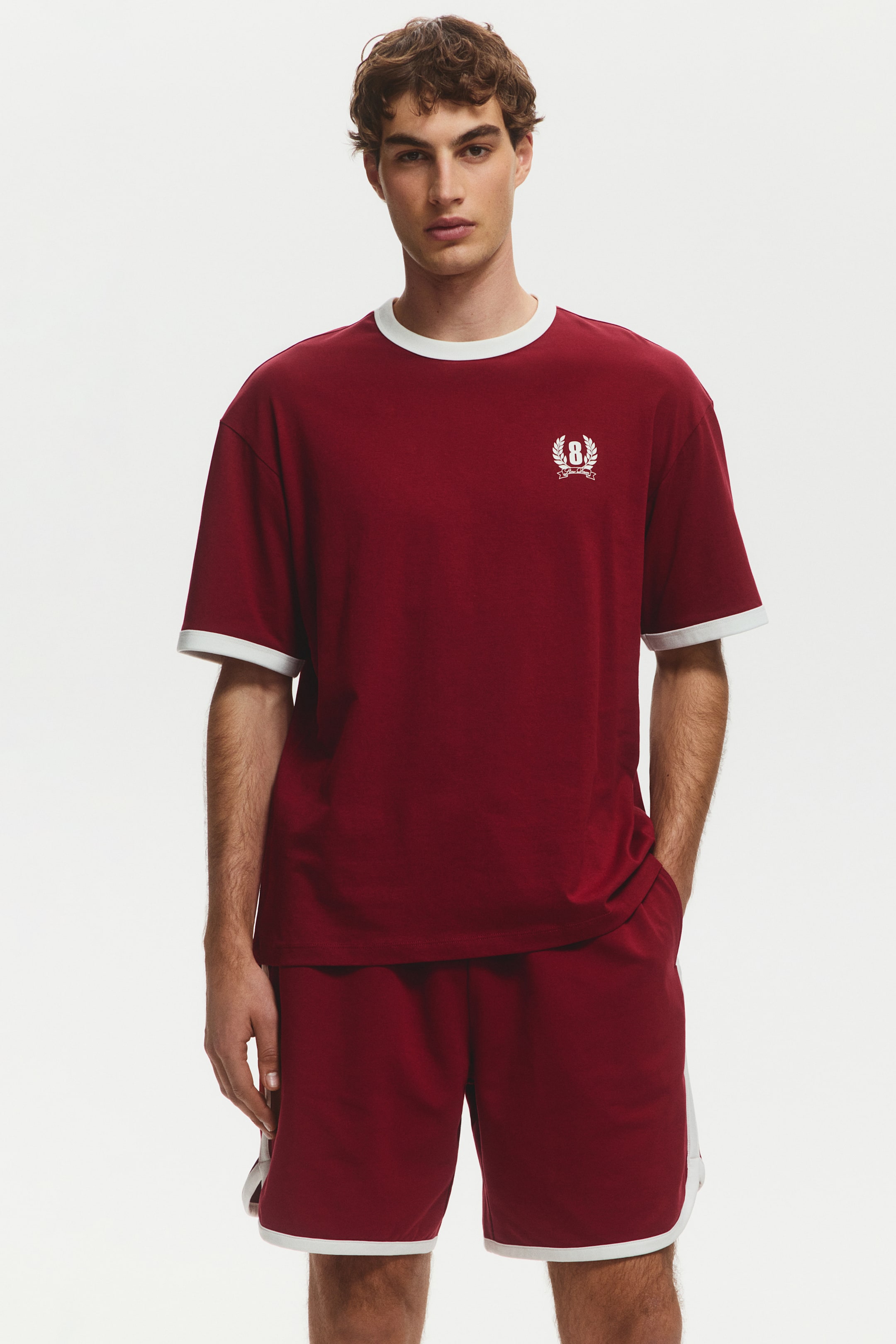 View larger image: A maroon t-shirt and shorts set is showcased from a front-facing angle. The t-shirt features a white ribbed crew neck, white ribbed short sleeve cuffs, and a white embroidered crest with the number '8' on the left chest. The matching shorts display white trim along the side seams and hem.