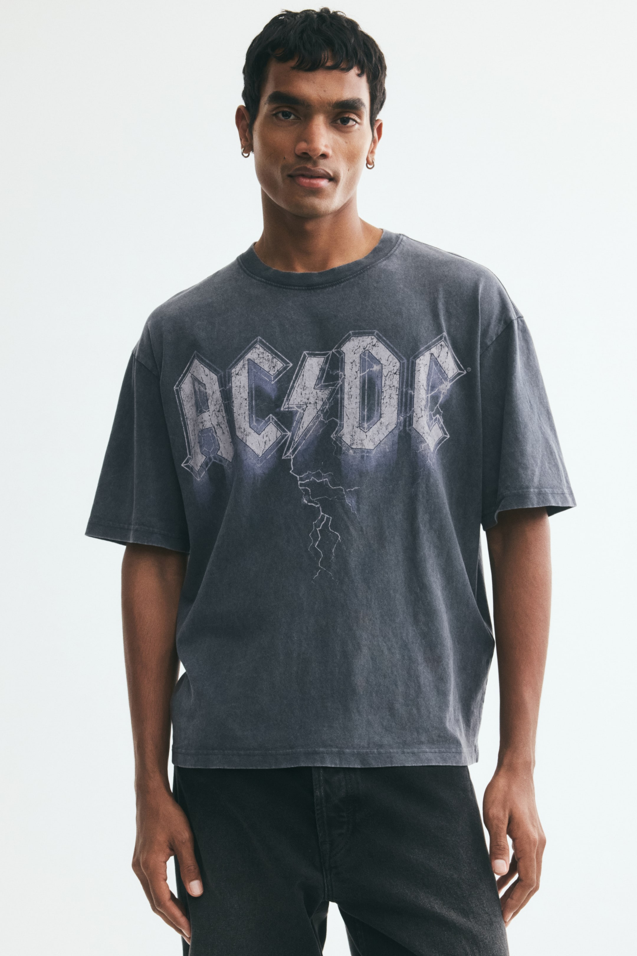 Zobacz większy obraz: Model ma na sobie ciemnoszary T-shirt z wyblakłym nadrukiem AC/DC. Koszulka ma okrągły dekolt wykończony ściągaczem, obniżoną linię ramion, prosty dół i swobodny krój, a stylizowana jest z czarnymi jeansami.