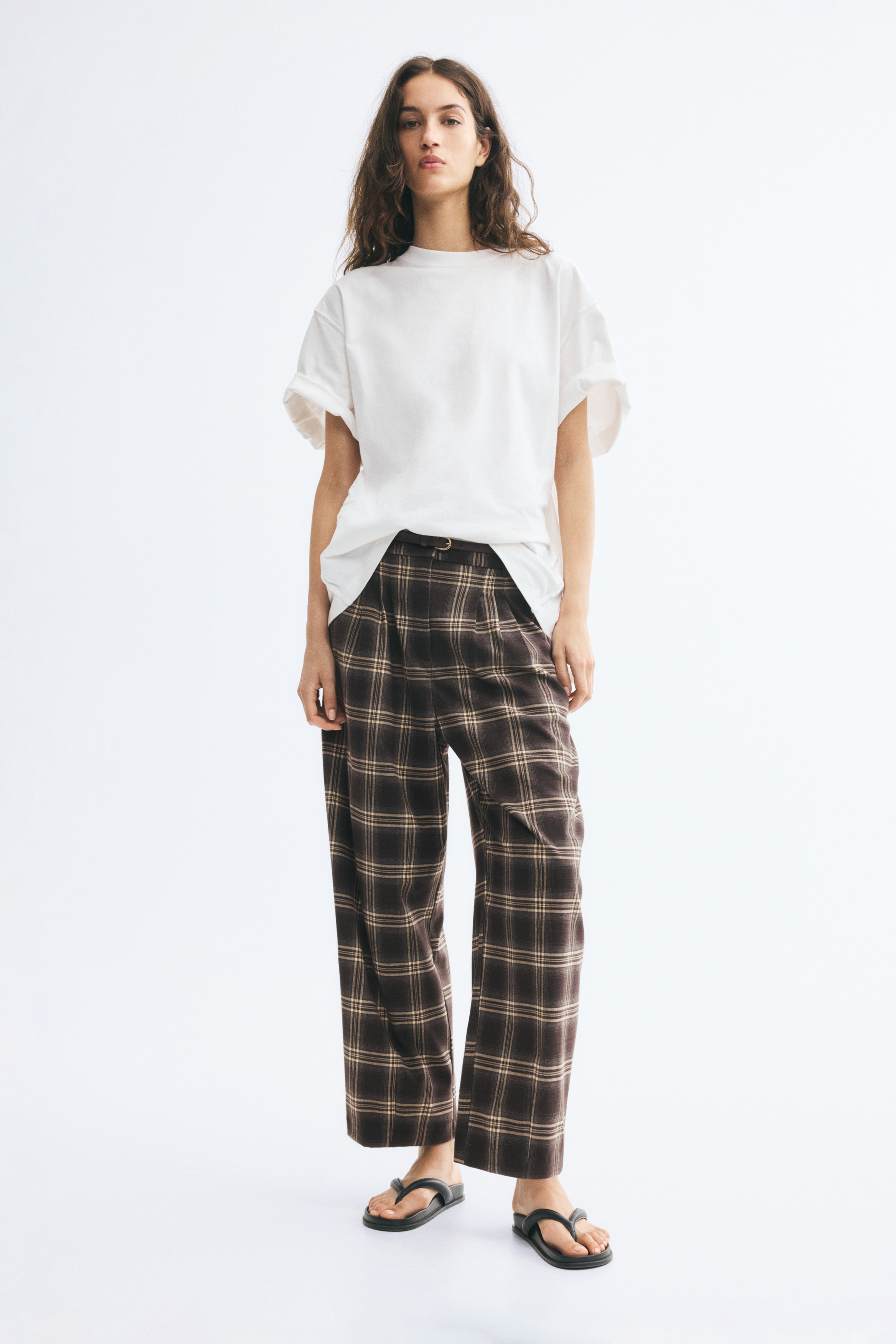 High-waisted, wide-leg trousers showcase a brown and beige plaid pattern from the front, featuring front pleats and a thin dark brown belt. The trousers hang straight, highlighting their structured waistband and relaxed silhouette.