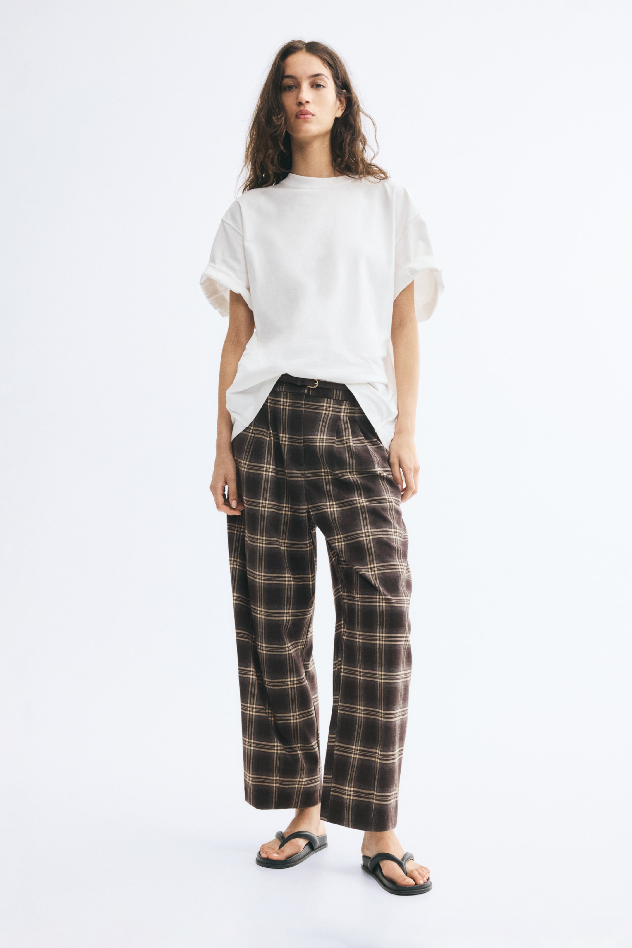 View larger image: Plaid, loose-fit, barrel-leg pants featuring front pleats and a high waist with a thin belt are worn by the model. The ankle-length pants are styled with a partially tucked-in white t-shirt and black thong sandals.