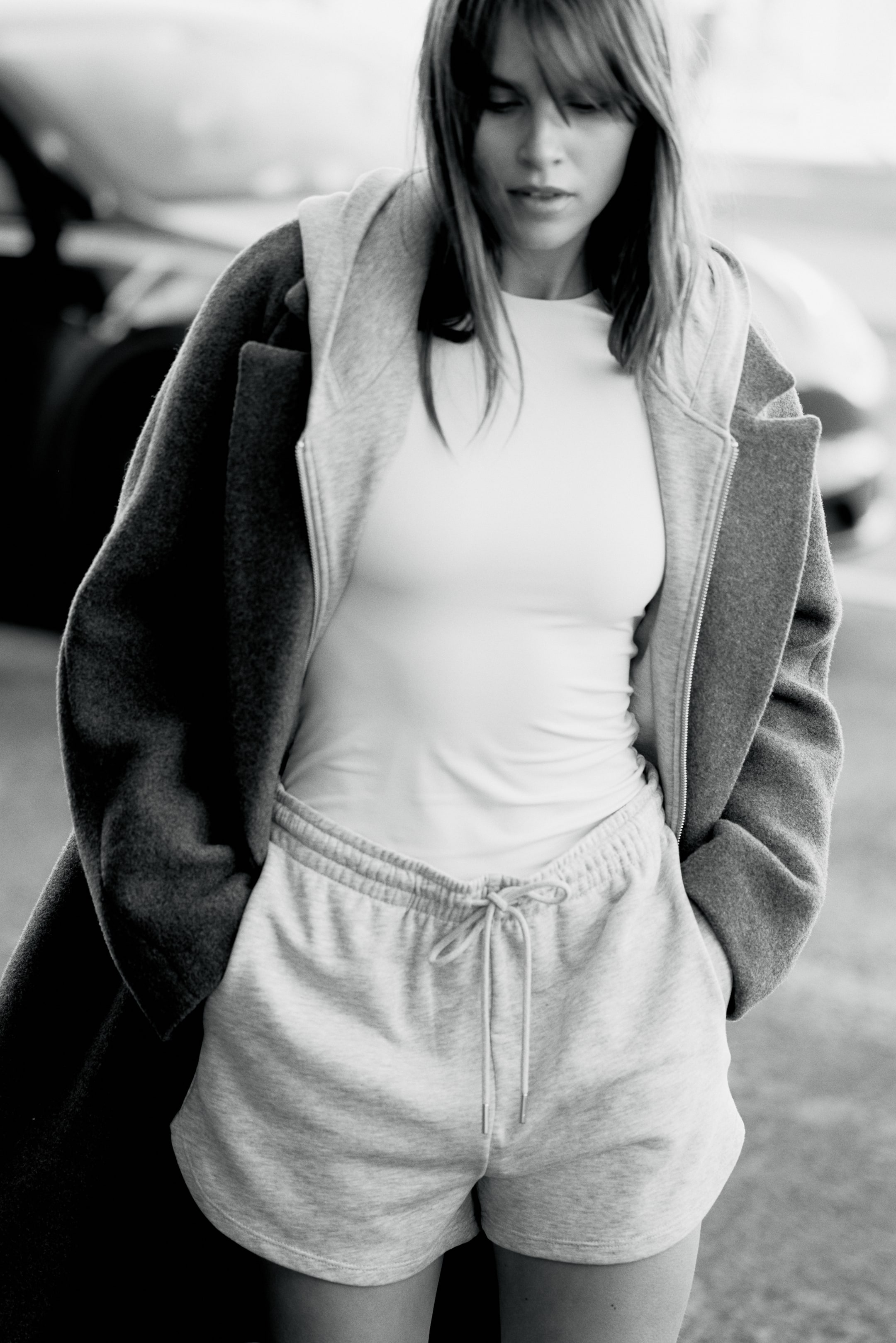 Büyük resmi görüntüle: Beyaz, vücuda oturan bir üst, açık gri fermuarlı kapüşonlu bir hırkanın altında giyiliyor ve bu hırkanın üzerinde koyu gri, bol kesim bir palto var. Açık gri polar şort, görünen bağları olan lastikli bir kordon bel ile dikkat çekiyor ve ayrıca giyiliyor.