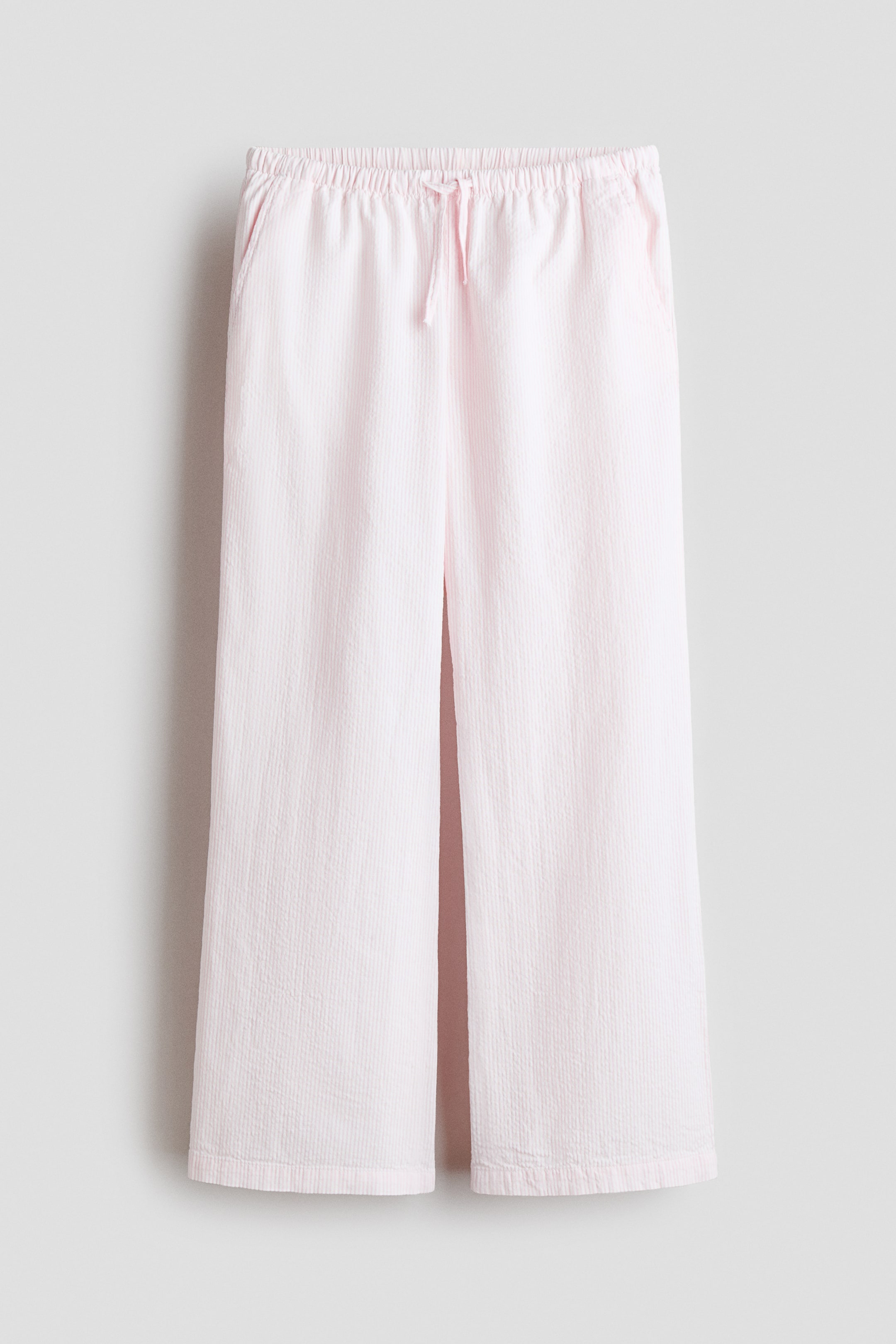 View larger image: Relaxed-fit light pink pants are shown from the front. They feature an elastic drawstring waistband with a visible tie, diagonal side pockets, and a woven fabric with a subtle vertical texture.