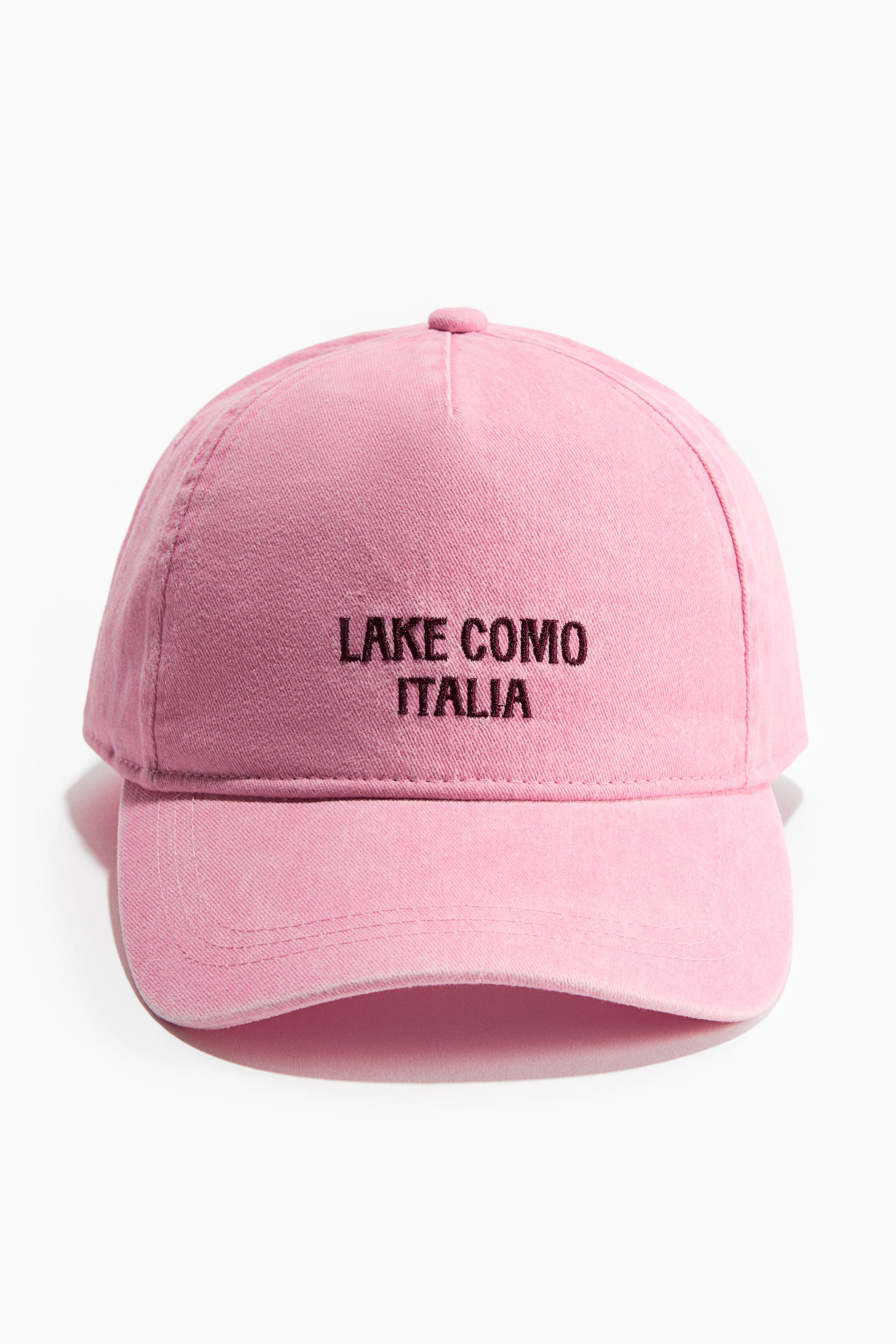 Ver imagen más grande: Una gorra de béisbol de algodón rosa lavado está orientada hacia el frente, mostrando "LAKE COMO ITALIA" bordado en hilo marrón oscuro en la parte delantera de la corona. Presenta una visera curvada con múltiples líneas de costura y un botón cubierto de tela en la parte superior de la corona.