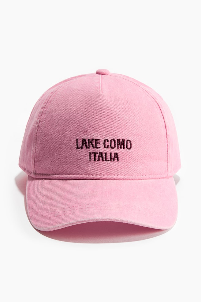 Une casquette de baseball en coton rose délavé est orientée vers l'avant, affichant "LAKE COMO ITALIA" brodé en fil marron foncé sur le devant de la couronne. Elle présente une visière incurvée avec plusieurs lignes de couture et un bouton recouvert de tissu sur le dessus de la couronne. - 1