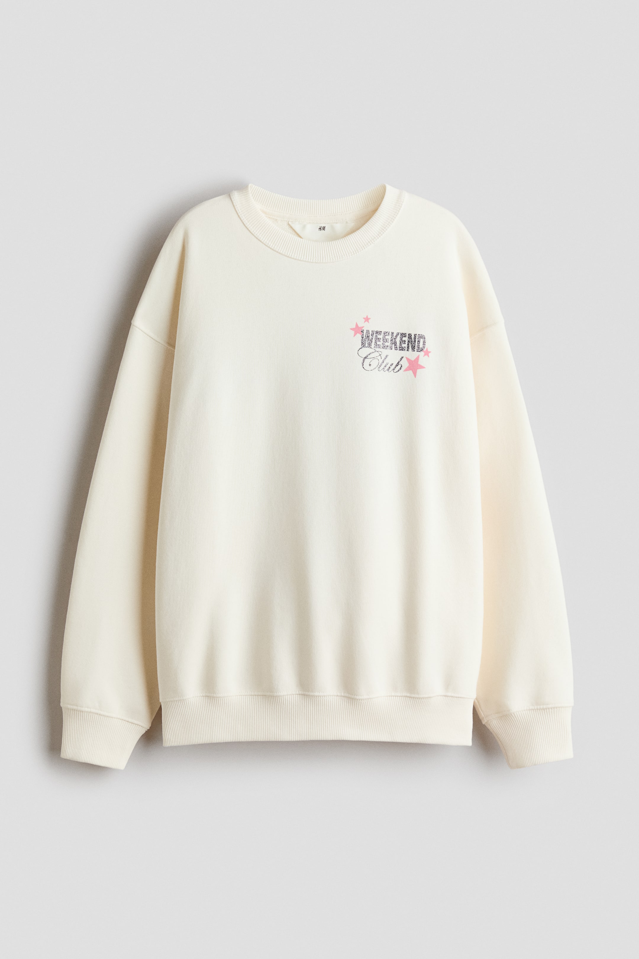Größeres Bild anzeigen: Ein locker sitzendes Sweatshirt in Creme wird von vorne gezeigt, mit einem glitzernden "WEEKEND Club"-Motiv und rosa Sternen auf der Brust. Es verfügt über einen runden, gerippten Halsausschnitt, überschnittene Schultern und lange Ärmel, wobei die Rippung die Bündchen und den Saum abschließt.