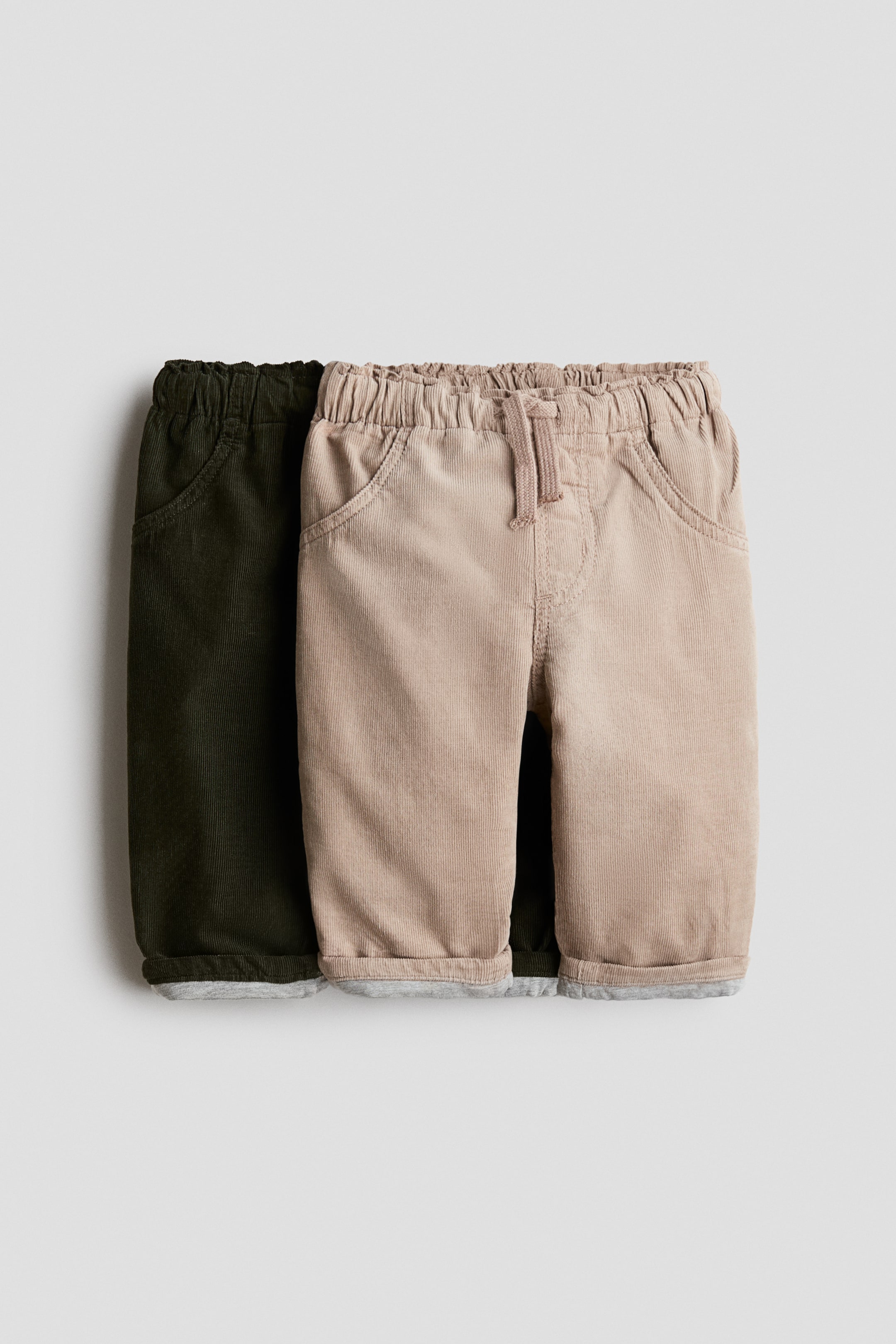 View larger image: Two pairs of baby corduroy pants are presented side by side, facing front. The beige pair features an elasticated drawstring waistband and slanted side pockets, with cuffed hems revealing a grey lining. The olive green pair is partially visible behind, showcasing the same design.