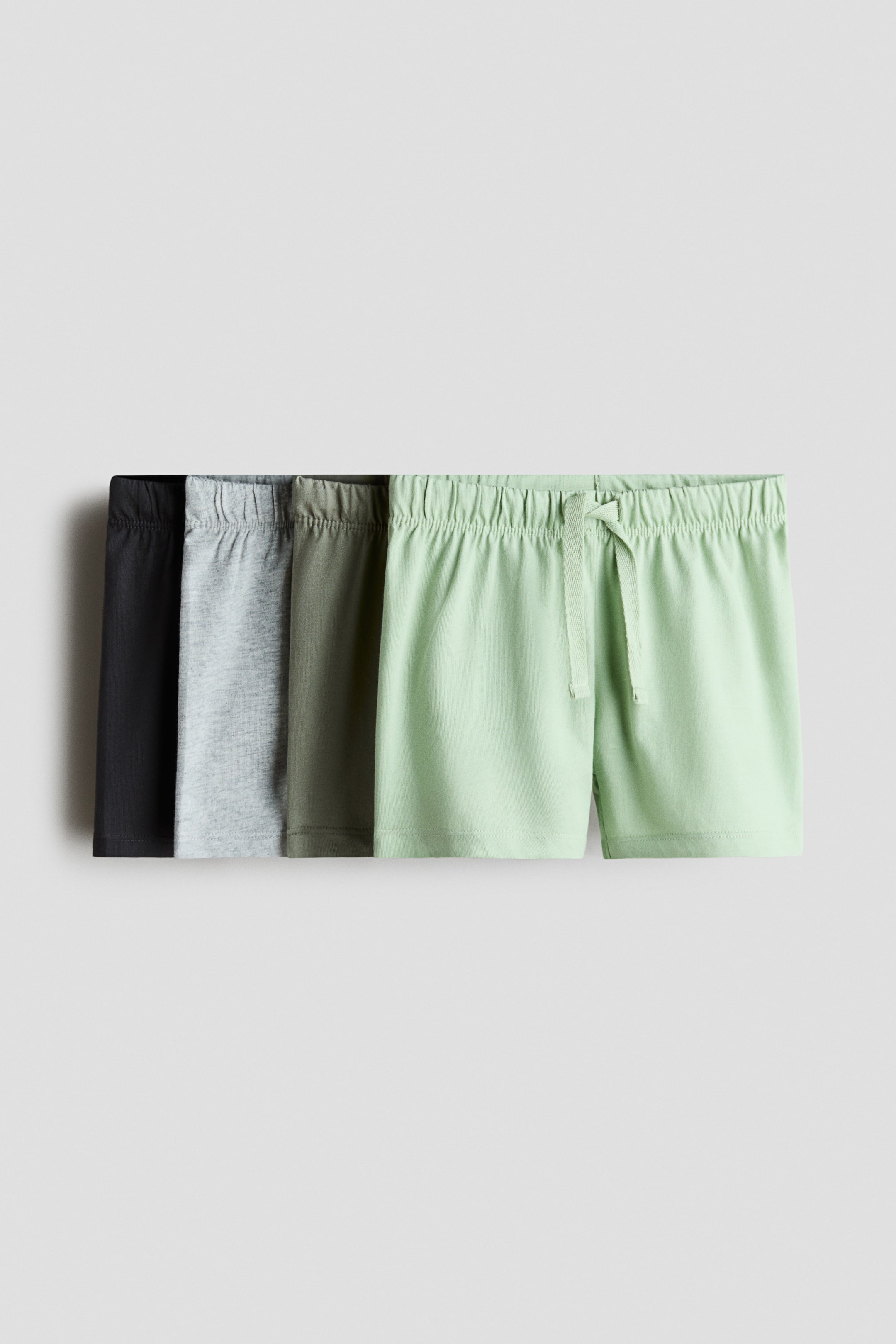 Visualizza immagine più grande: Quattro paia di morbidi pantaloncini in jersey di cotone sono mostrati frontalmente, impilati orizzontalmente, ciascuno dotato di vita elasticizzata. Il paio verde chiaro in primo piano presenta anche un cordino visibile, mentre gli altri pantaloncini appaiono in nero, grigio chiaro e verde oliva.