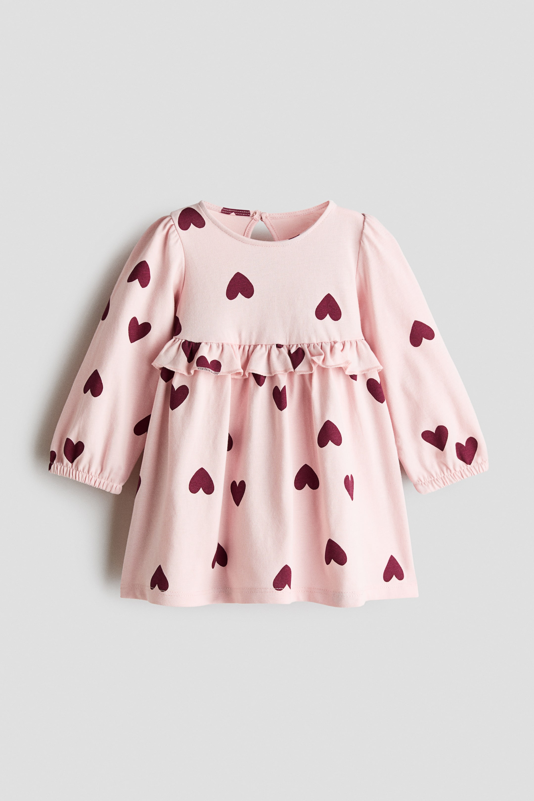 View larger image: A baby frock showcases a soft pink hue adorned with a pattern of deep red hearts, front-facing. It boasts a rounded neckline with a keyhole and button fastening at the back, long sleeves with elasticised cuffs, and a ruffled accent across the front bodice.