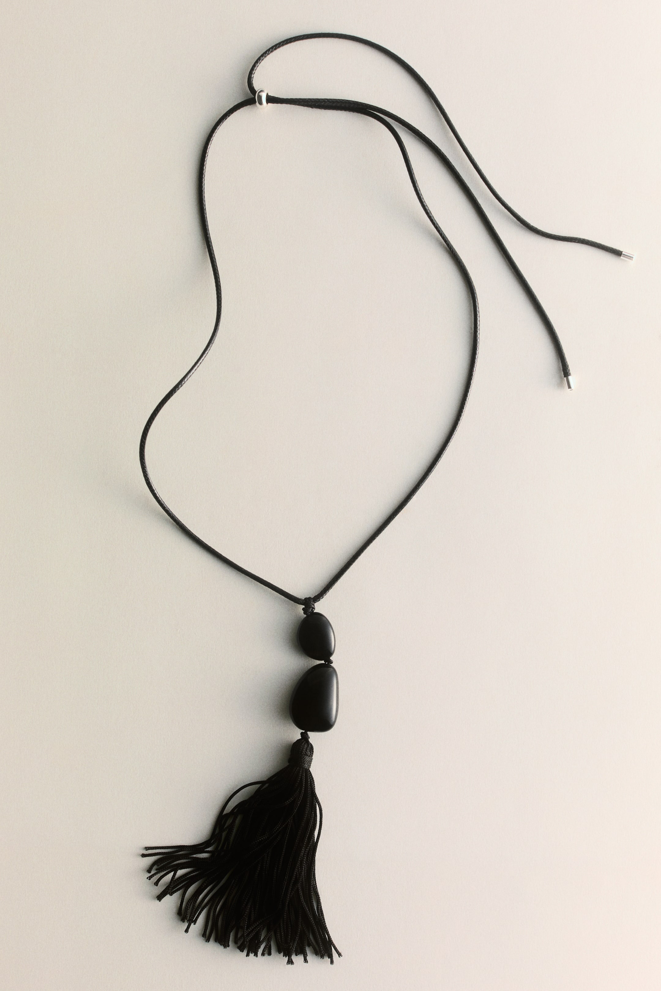 Grotere afbeelding bekijken: Een zwarte koordketting ligt plat en heeft een verstelbare zilverkleurige schuifregelaar op het koord. Twee gladde, onregelmatig gevormde zwarte kralen vormen een hanger, met daaronder een zwarte kwast.