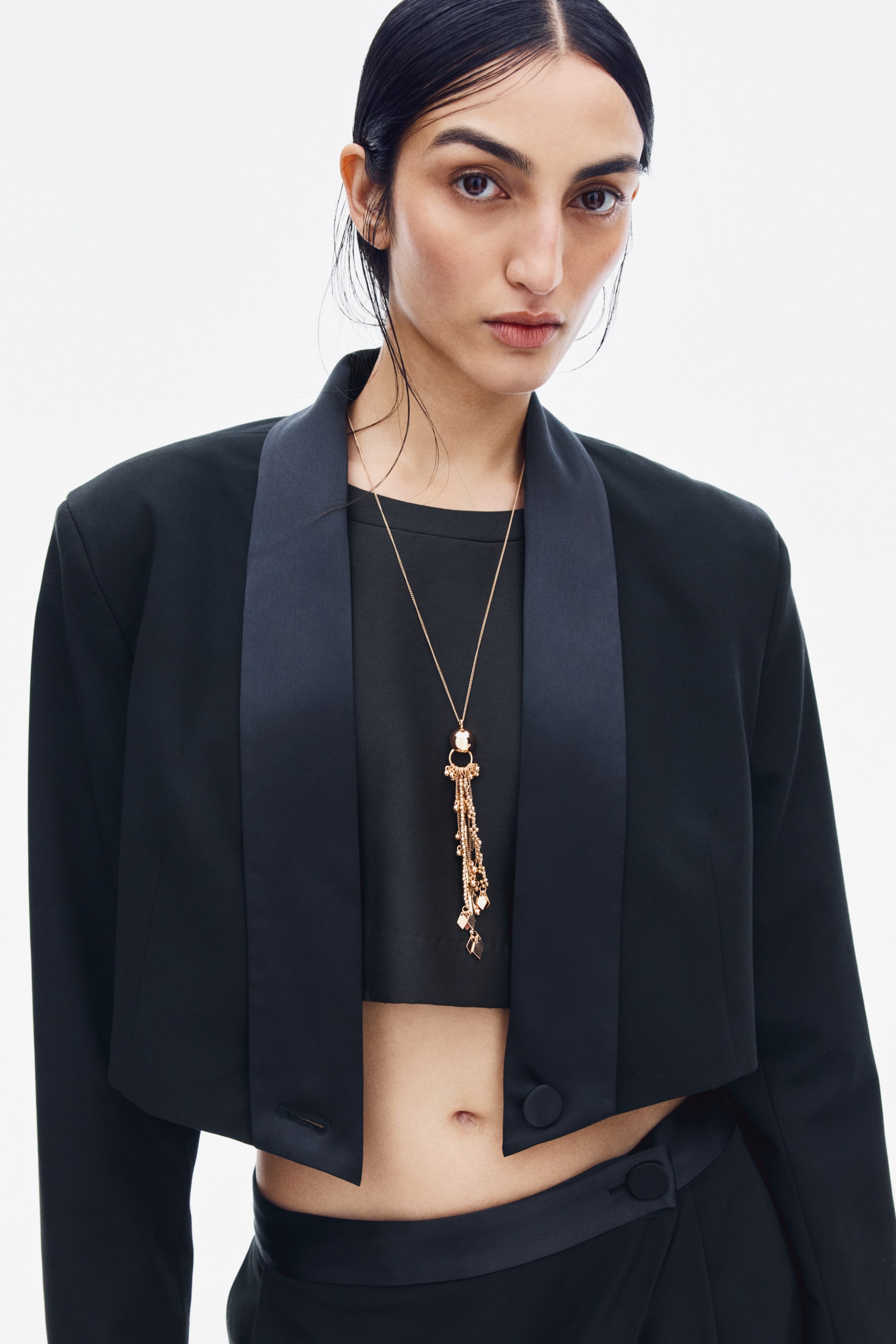 View larger image: A long metal chain necklace featuring a gold-toned spherical bead and a multi-strand chain tassel is being worn by the model. The necklace hangs over a black cropped top layered under a matching open cropped jacket.