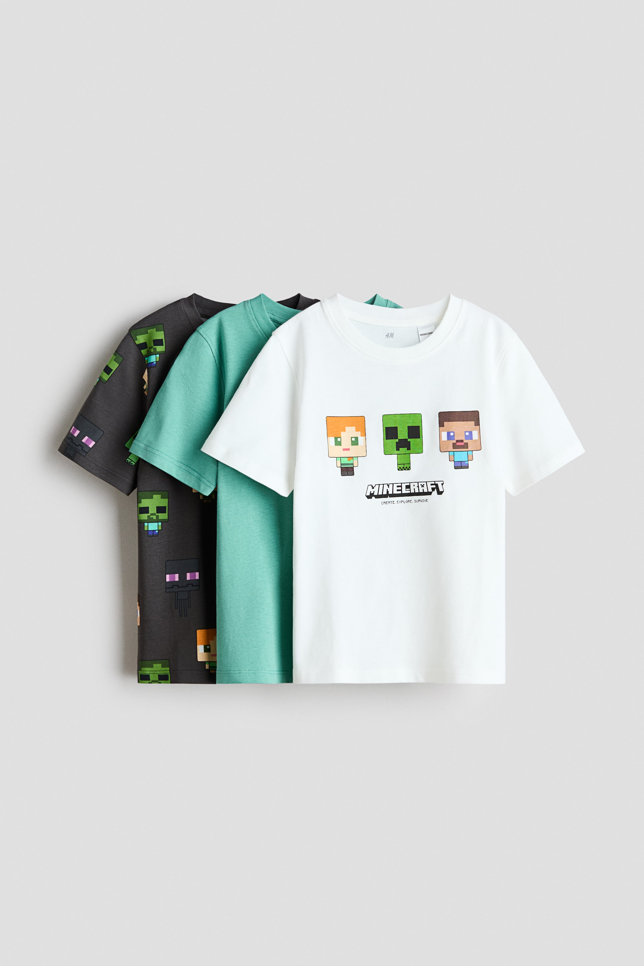 Agrandir l'image: Trois t-shirts sont superposés, avec le t-shirt blanc à l'avant affichant un imprimé graphique de trois personnages pixélisés et le texte "MINECRAFT", et doté d'un col à bord-côte. Un t-shirt turquoise est superposé derrière, et un t-shirt gris foncé avec un imprimé intégral de personnages pixélisés est superposé derrière le turquoise.