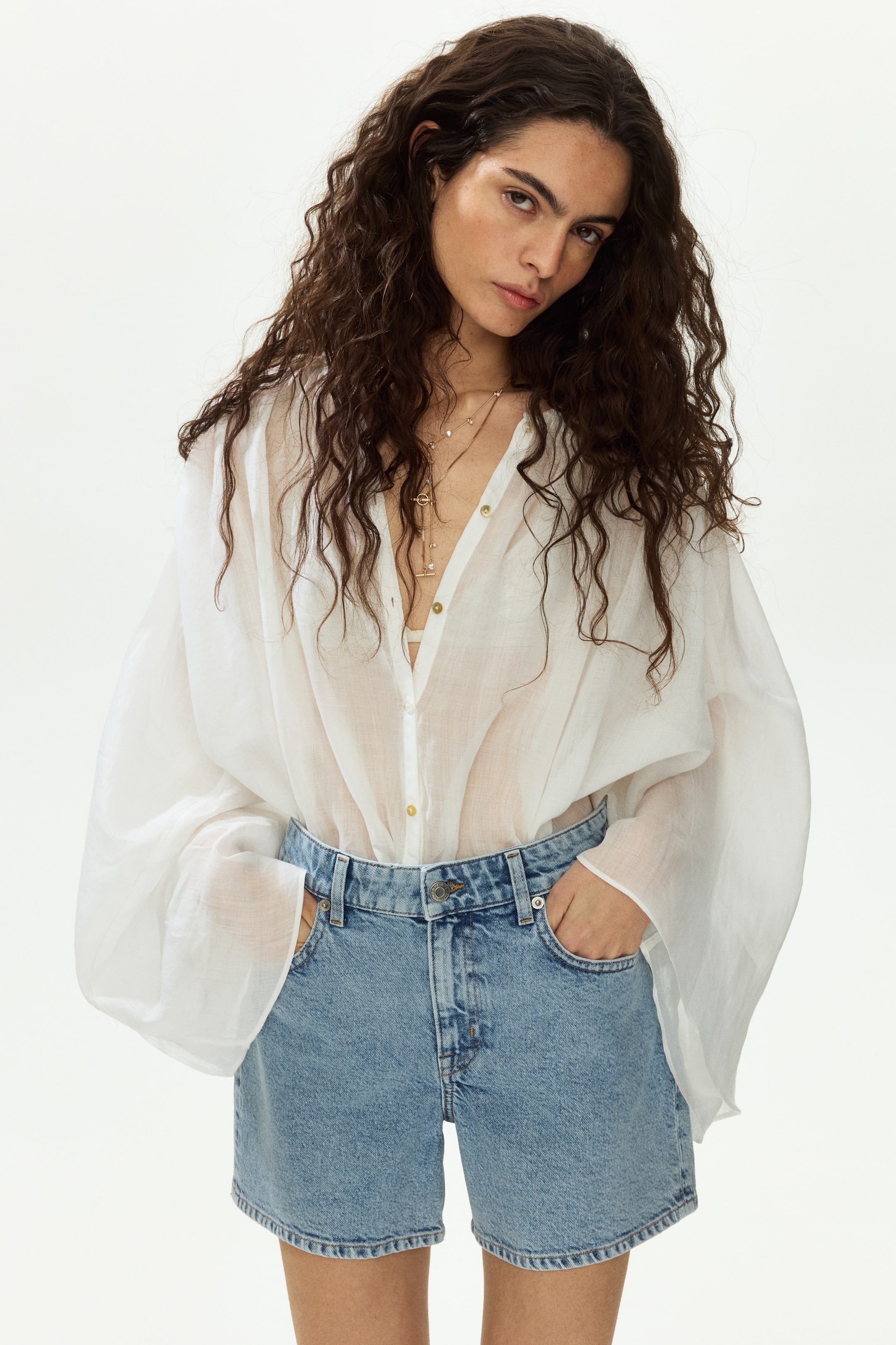 Xem ảnh lớn hơn: Một chiếc áo blouse trắng mỏng, có cúc cài phía trước, đang được mặc, với chất liệu vải lộ ra chiếc áo lót màu sáng và có tay áo dài, phồng, cùng với các nút cài màu vàng ở phía trước. Chiếc áo blouse được sơ vin một phần vào quần short denim cạp cao, màu sáng, có kiểu dáng thoải mái và túi phía trước.