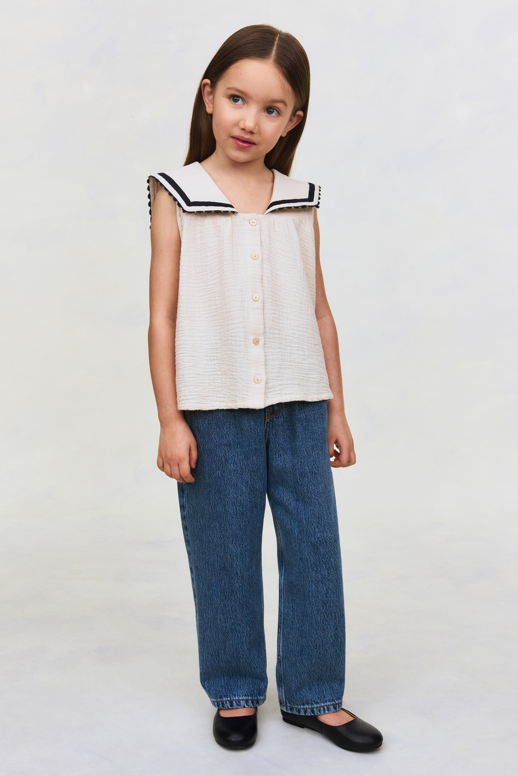 View larger image: A sleeveless, light-beige textured top featuring a cream sailor collar with black trim and bobble detailing, with buttons down the front and paired with loose-fitting, mid-blue denim jeans.