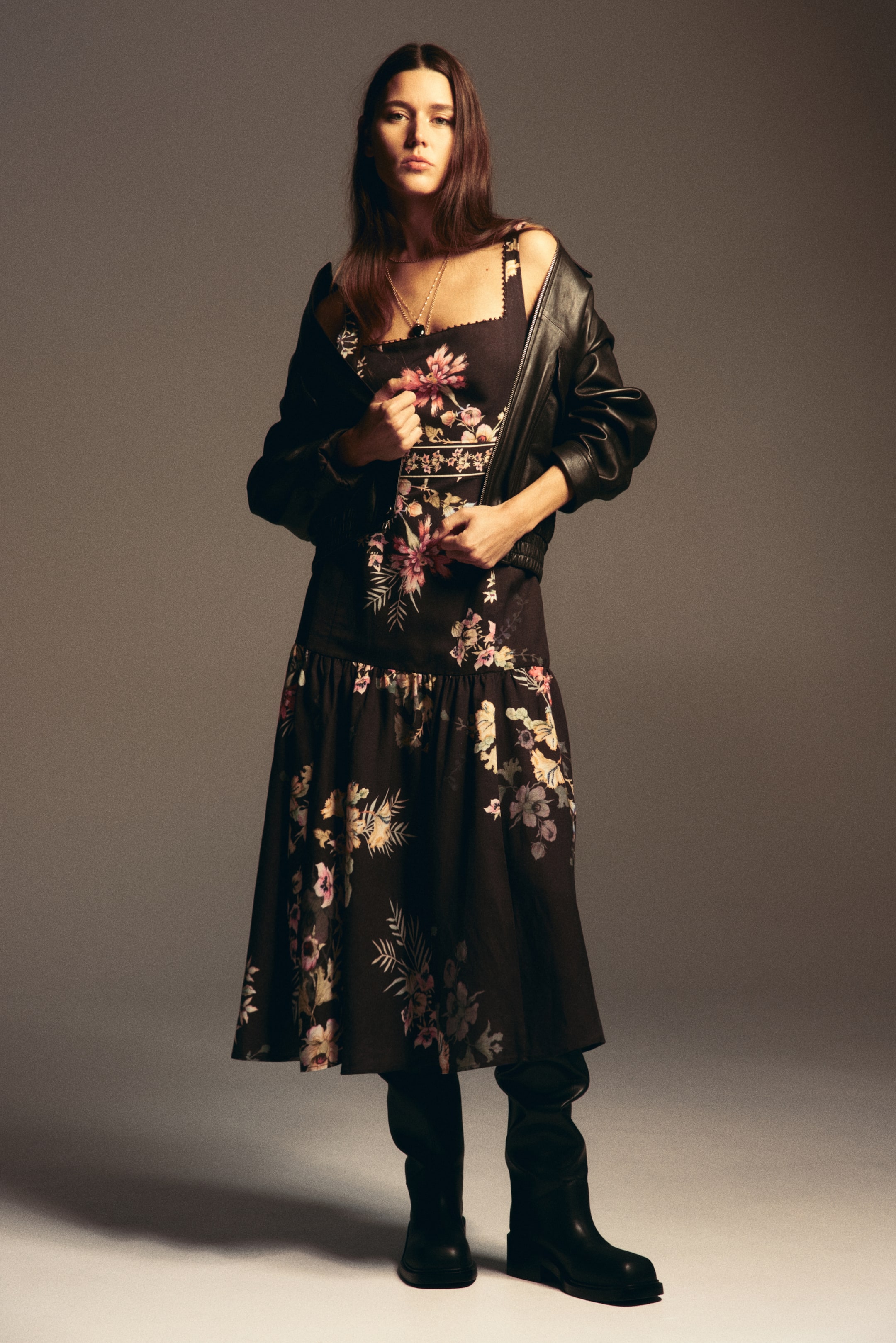 Prikaži veću sliku: Tamna cvetna midi haljina ima četvrtasti izrez sa talasastim rubom, tanke bretele, horizontalni šareni pojas na telu haljine i nabrani spušten struk. Preko haljine je slojevit crni kožni bomber jakna, prebačena preko ramena, a ispod poruba vidljive su crne visoke čizme.