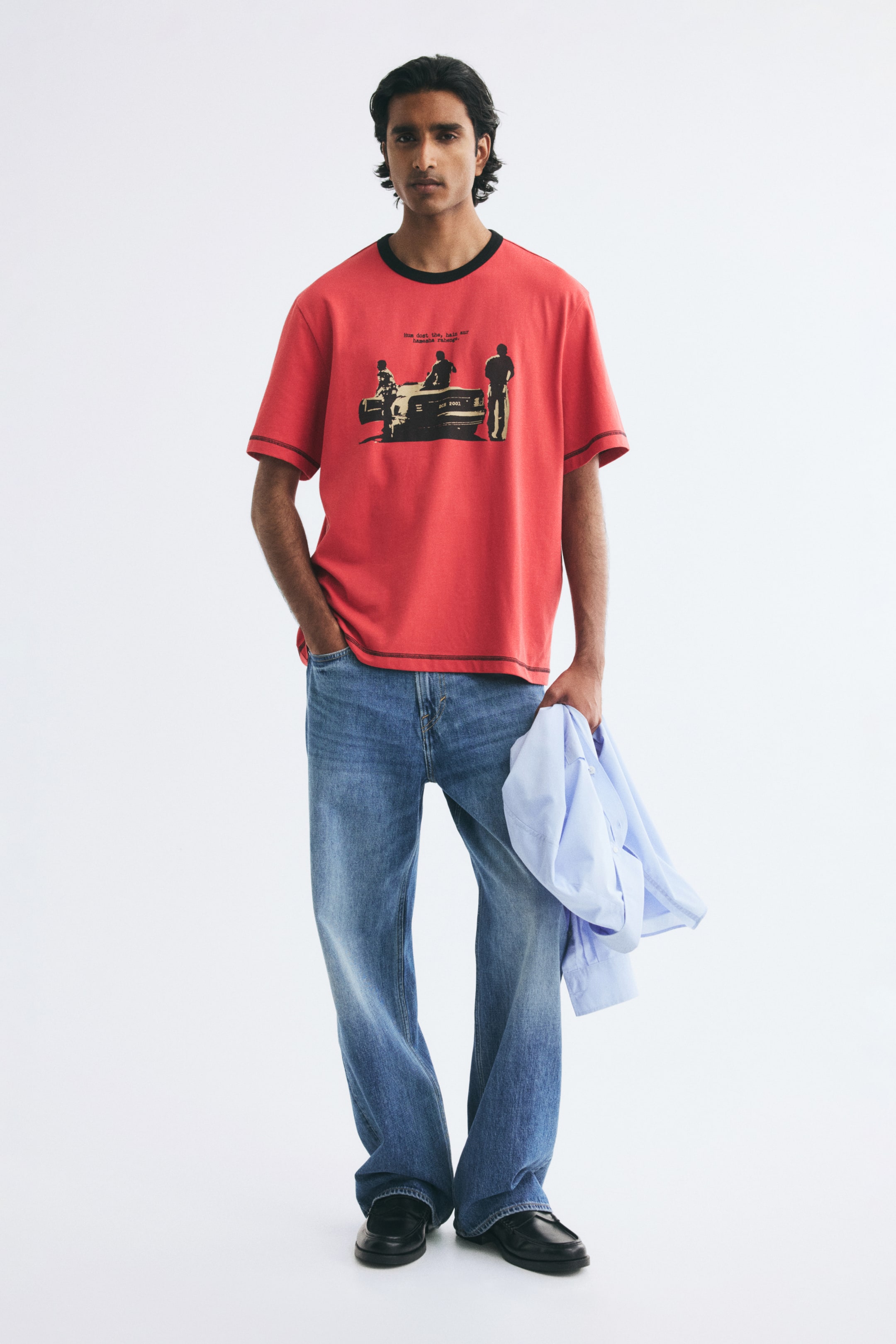 View larger image: A red, regular-fit T-shirt featuring a black graphic print of figures near a car is worn by the model. The T-shirt is styled with a black ribbed neckline, gently dropped shoulders, and visible dark flatlock stitching along the cuffs and hem. The model pairs the tee with blue jeans while holding a light blue long-sleeve shirt in one hand.