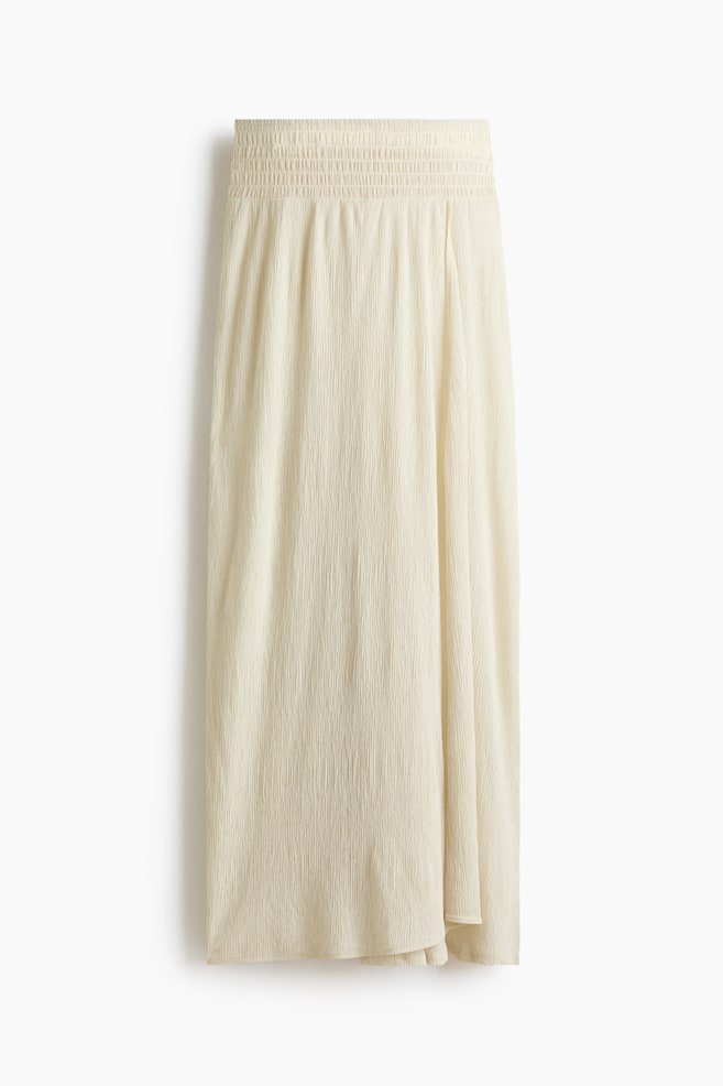 A cream-coloured skirt is laid flat, featuring a shirred, elasticated waistband and a crinkled texture throughout the fabric. - 1