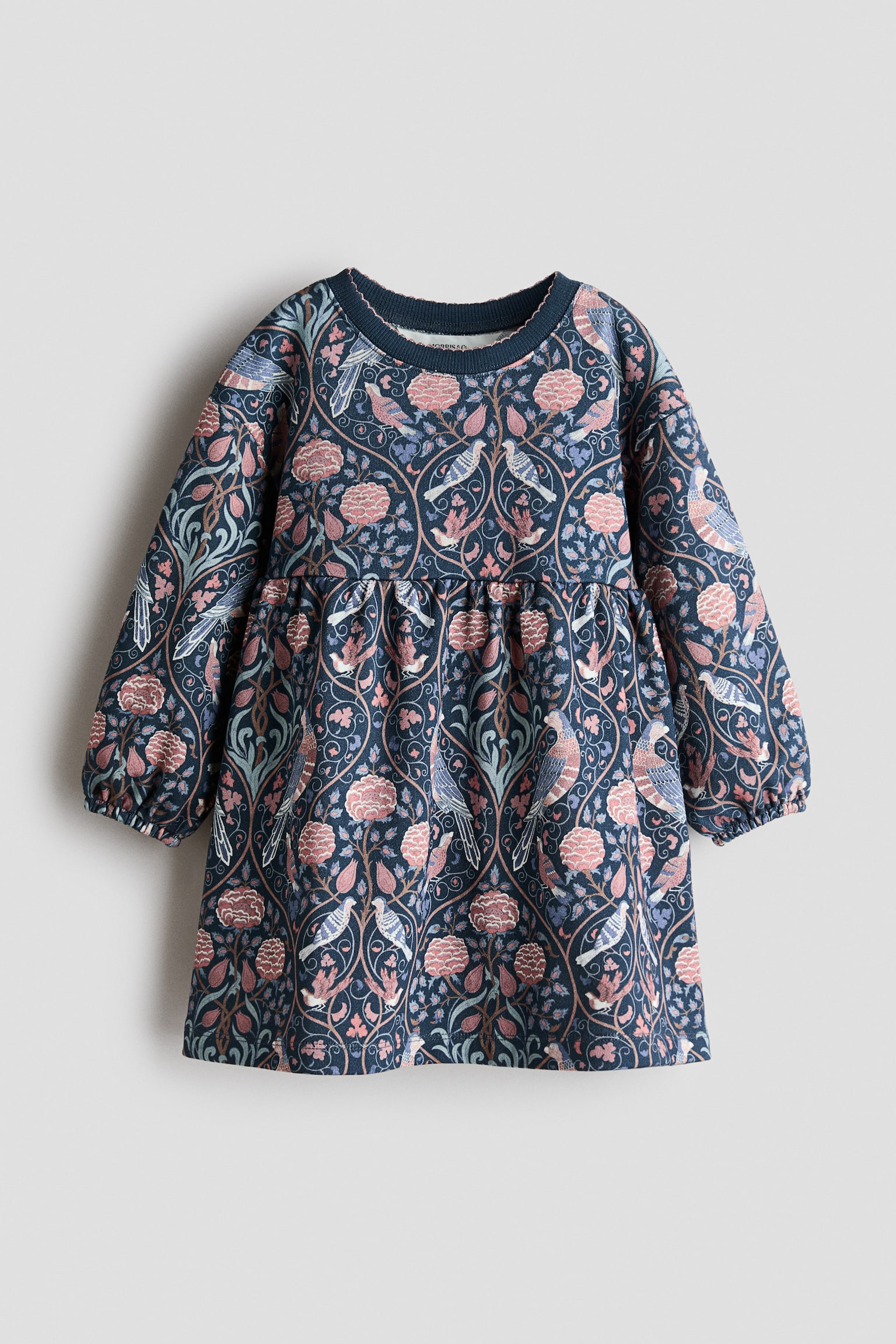 Ein langärmliges Kleid in Frontansicht zeigt ein Allover-Muster aus Vögeln und Blumen in Rosa-, Weiss- und Blautönen auf dunkelblauem Hintergrund. Das Design verfügt über einen runden, mit Rippstrick eingefassten Ausschnitt mit zarter Spitzenborte, überschnittene Schultern, voluminöse Ãrmel mit elastischen Bündchen sowie eine Raffung in der Taille, die in einen ausgestellten Rock übergeht. - 1