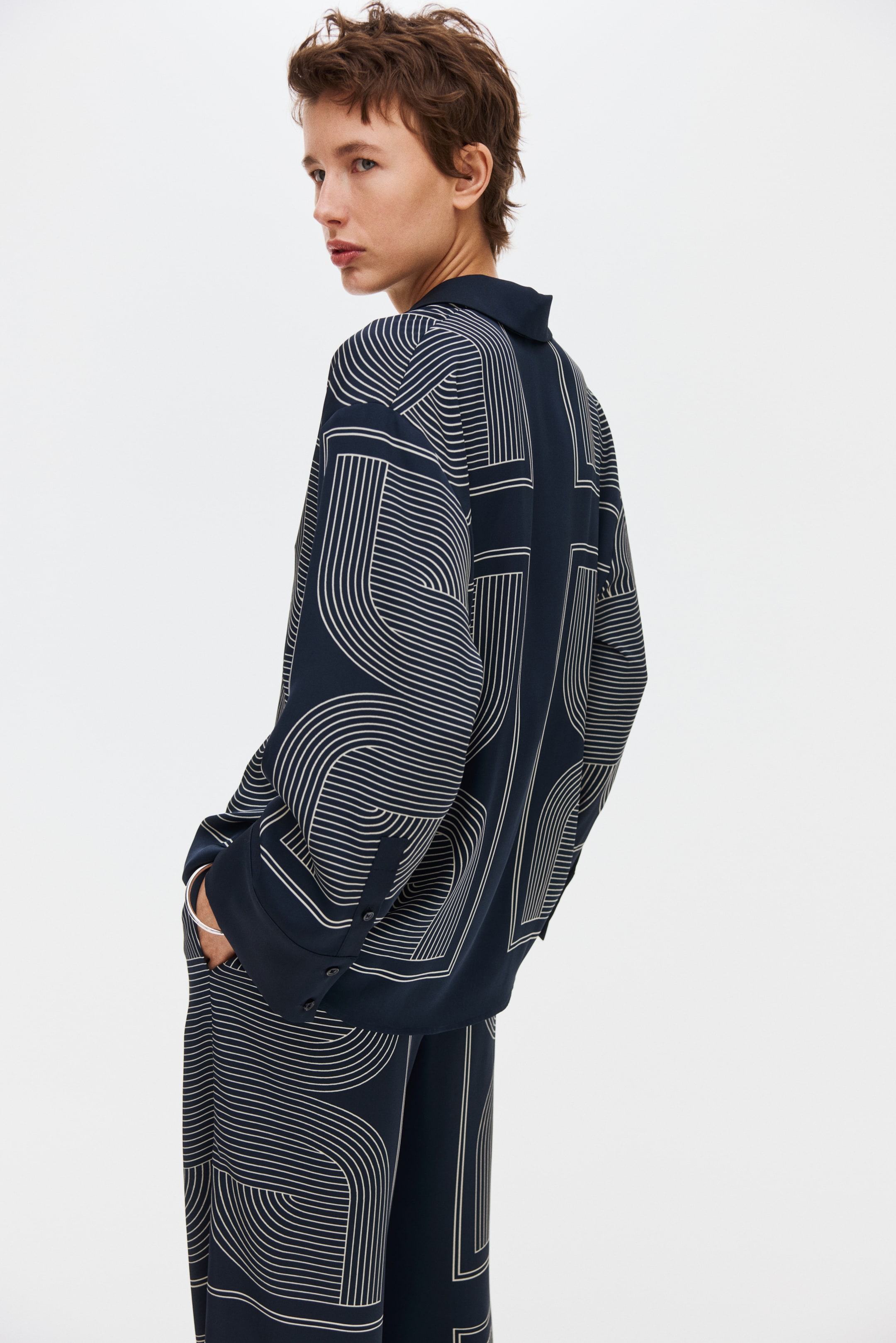 View larger image: A navy-blue long-sleeved shirt and matching wide-leg trousers feature a geometric pattern of light-coloured curved and straight lines. The loose-fitting shirt, shown from a back-side angle, has a collar and buttoned cuffs, with the pattern prominent across its back and sleeves, while the trousers continue the same design.