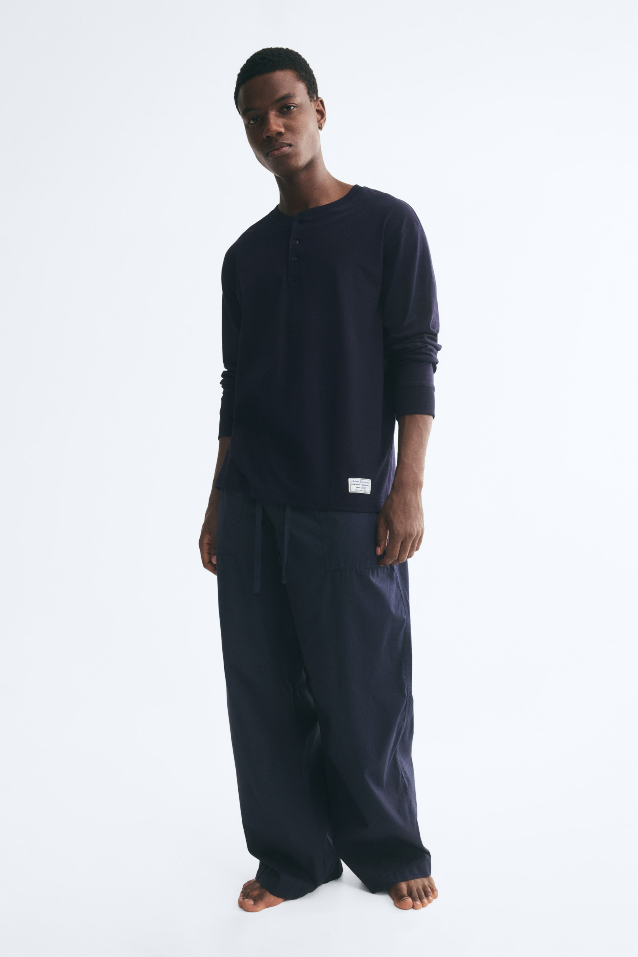 View larger image: A dark navy blue cotton loungewear set is being worn by the model, featuring a long-sleeve jersey top with a button placket and a small white tag near the hem. The matching wide-leg woven trousers feature an adjustable drawstring waist and side pockets.