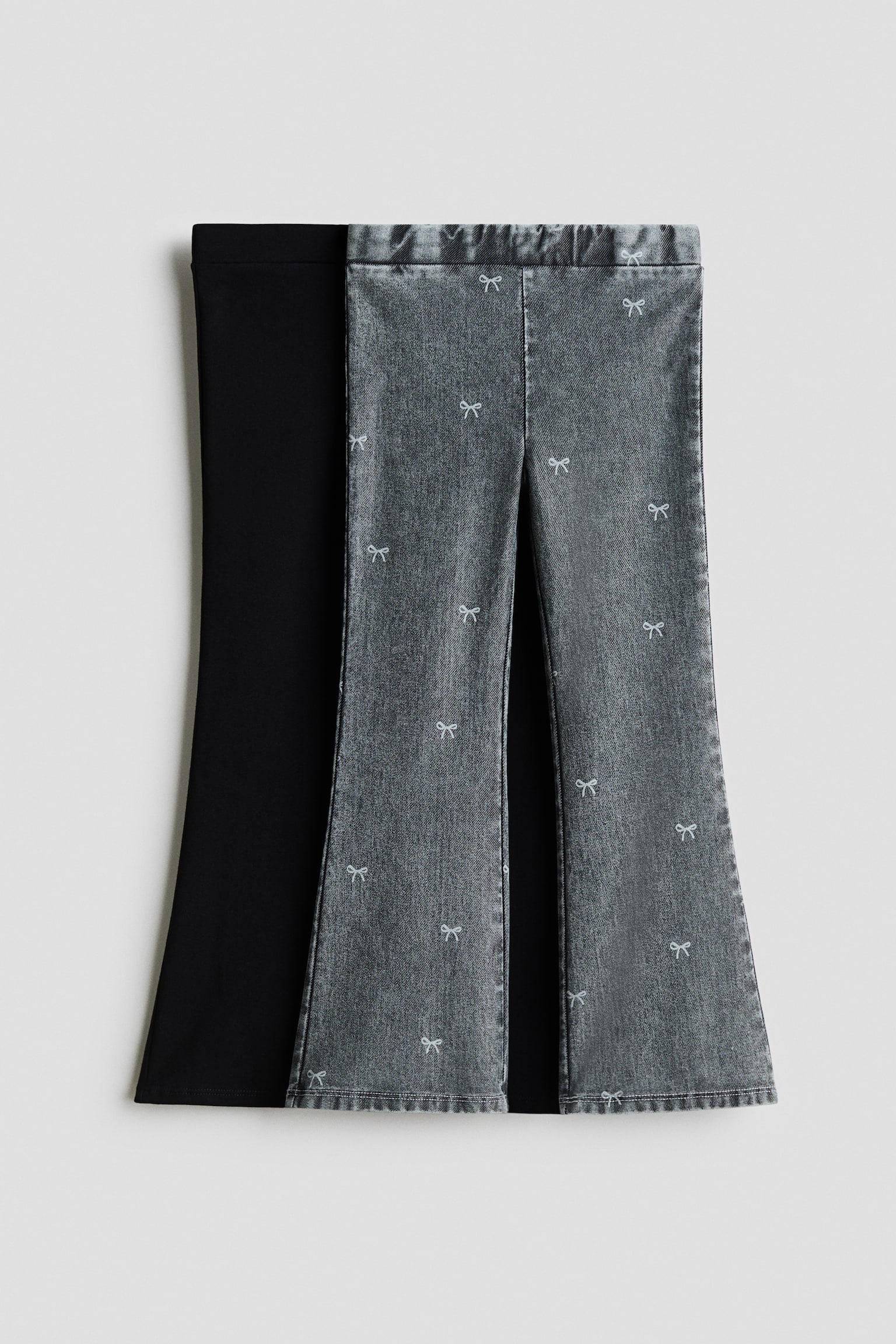 Two pairs of flared jeggings are shown from the front, featuring an easy pull-on waist and flared legs. One pair is displayed in grey, denim-look jersey with a scattered pattern of small white bows, while the overlapping pair behind it is shown in solid black jersey fabric.