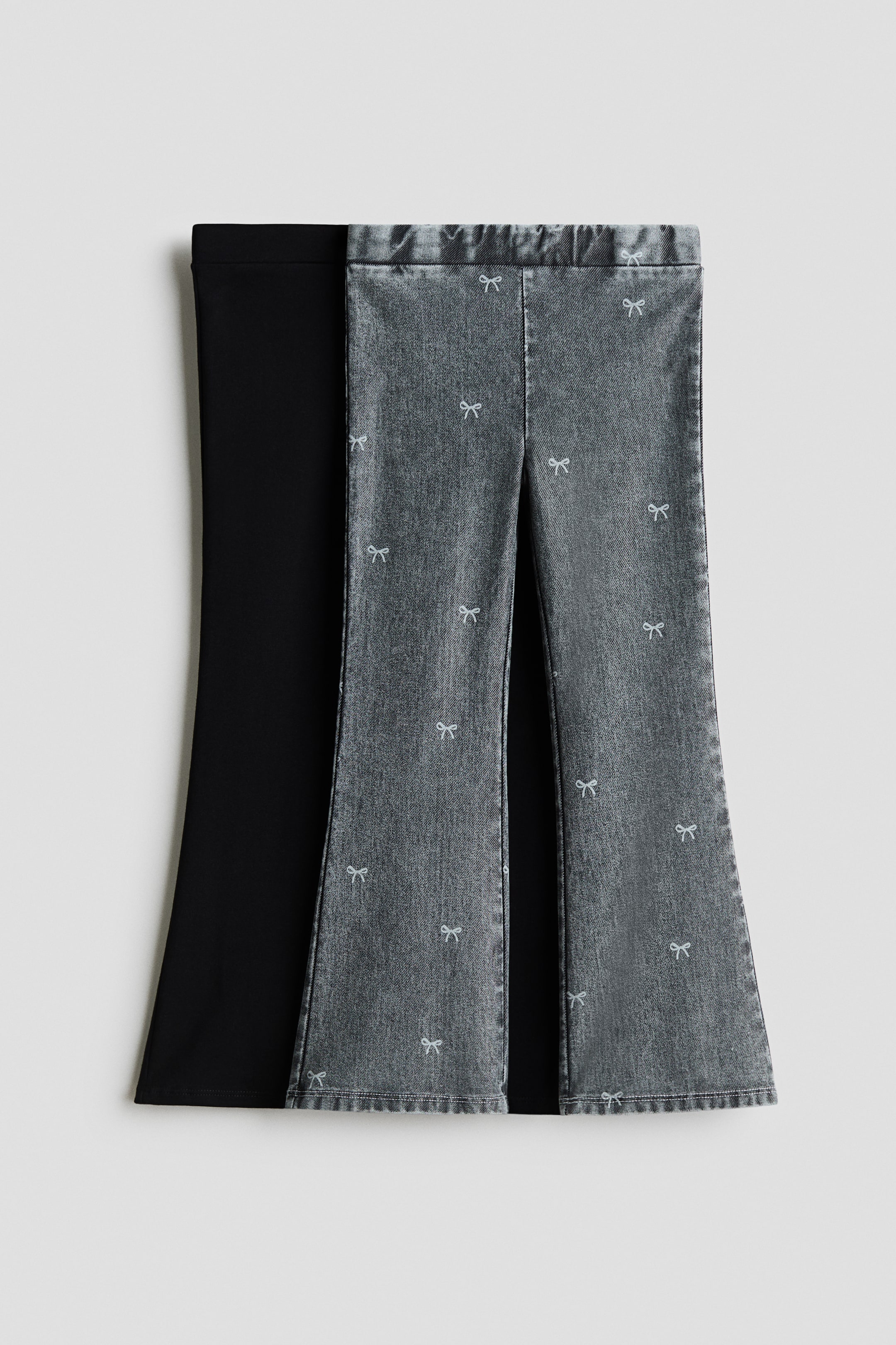 View larger image: Two pairs of flared jeggings are shown from the front, featuring an easy pull-on waist and flared legs. One pair is displayed in grey, denim-look jersey with a scattered pattern of small white bows, while the overlapping pair behind it is shown in solid black jersey fabric.