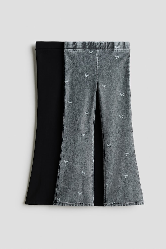 Two pairs of flared jeggings are shown from the front, featuring an easy pull-on waist and flared legs. One pair is displayed in grey, denim-look jersey with a scattered pattern of small white bows, while the overlapping pair behind it is shown in a solid black jersey fabric. - 1