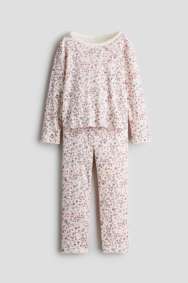 Un ensemble de pyjama est prÃ©sentÃ© de face, composÃ© dâun haut Ã  manches longues et dâun pantalon assorti en tissu de couleur crÃ¨me, avec un imprimÃ© floral violet et rose sur lâensemble. Le haut est dotÃ© dâun col rond, et les deux piÃ¨ces sont finies avec une dÃ©licate bordure picot au niveau de lâencolure, des poignets et des ourlets. - 1