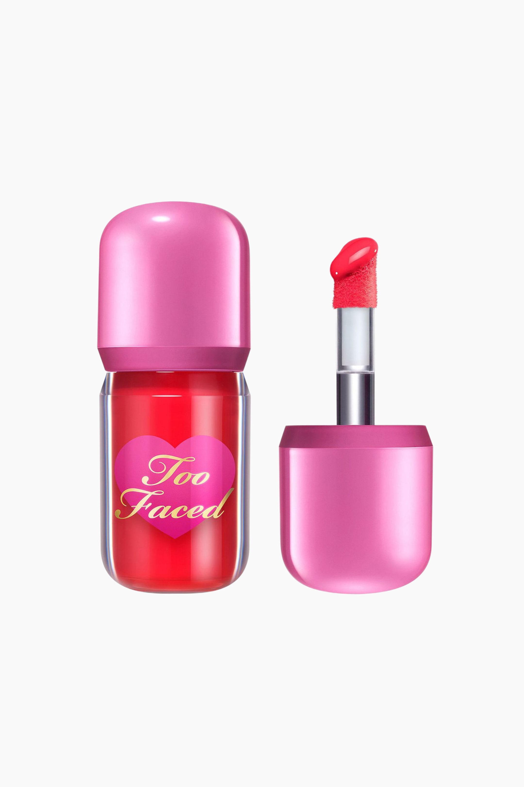 Visa större bild: En Too Faced läppfärg visas, bestående av en genomskinlig flaska fylld med livligt röd vätska, prydd med en rosa hjärtetikett med guldfärgad 'Too Faced' text och en rundad rosa kork. Bredvid den ligger den lösa applikatorn som visar en rosa flockad hjortfotspets täckt med röd produkt, som kommer från en silverstam och vilar i en inverterad rosa kork.