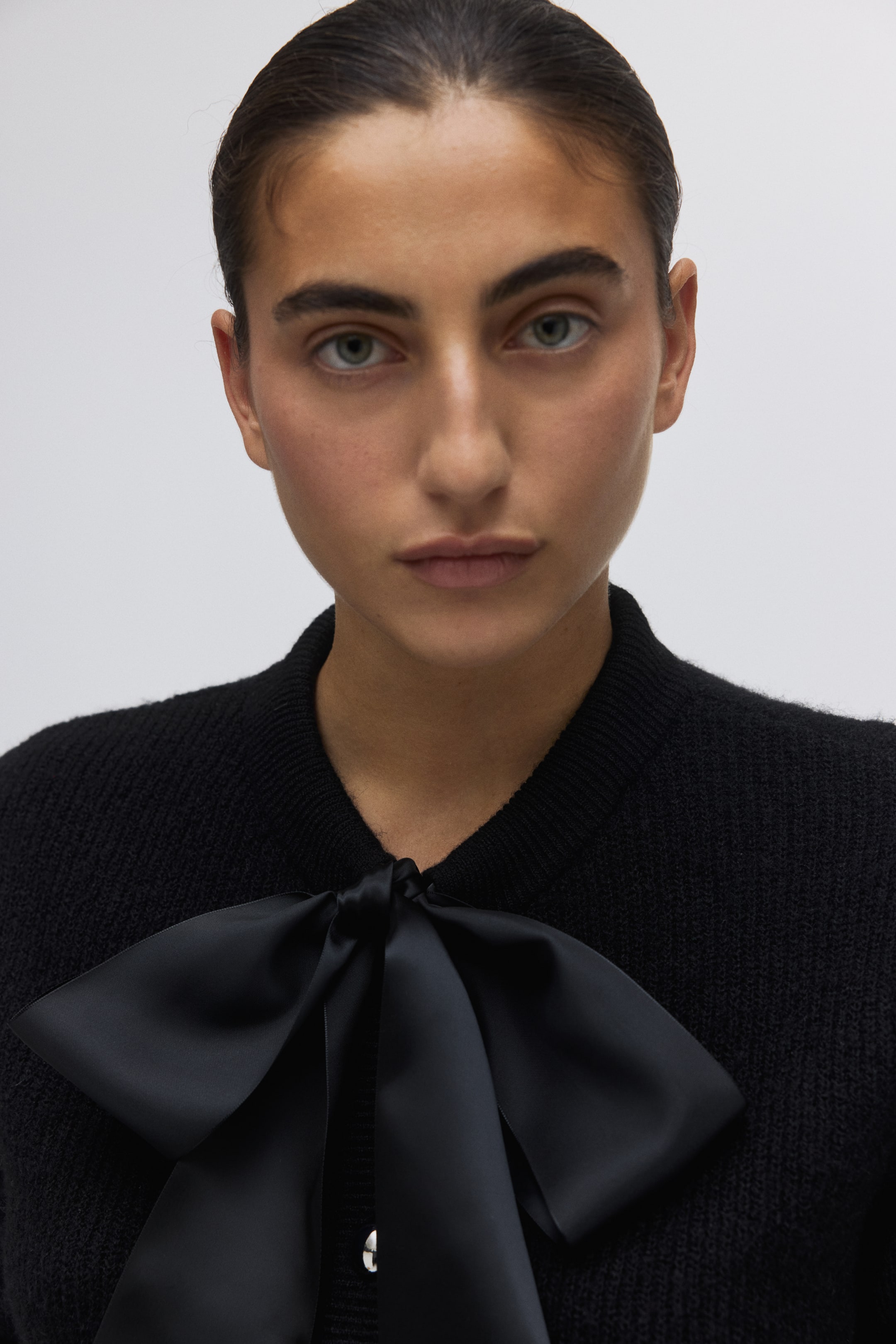 Grotere afbeelding bekijken: Een zwart gebreid vest heeft een opvallende zwarte satijnen strik die aan de voorkant is vastgebonden, gepositioneerd onder een ronde halslijn, met een enkele parelachtige knop zichtbaar op de sluiting.