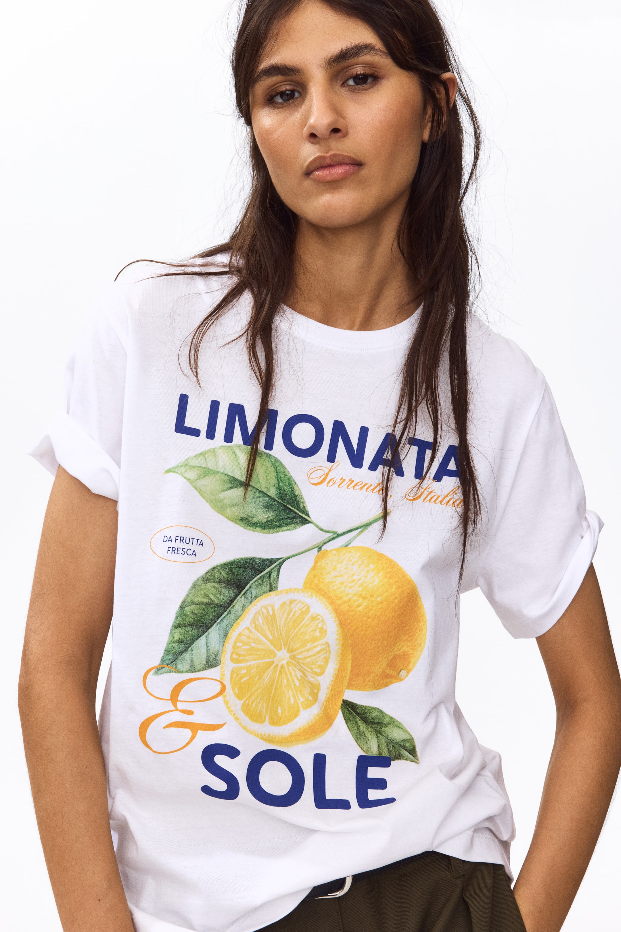 查看大圖: 白色T恤上展示了两个柠檬的图案，带有绿色的叶子和茎，一个完整的，一个切片，旁边是蓝色的文字“LIMONATA”和“E SOLE”，以及较小的橙色文字“Sorrento, Italia”和“DA FRUTTA FRESCA”。这款T恤采用卷起的短袖和圆领设计。