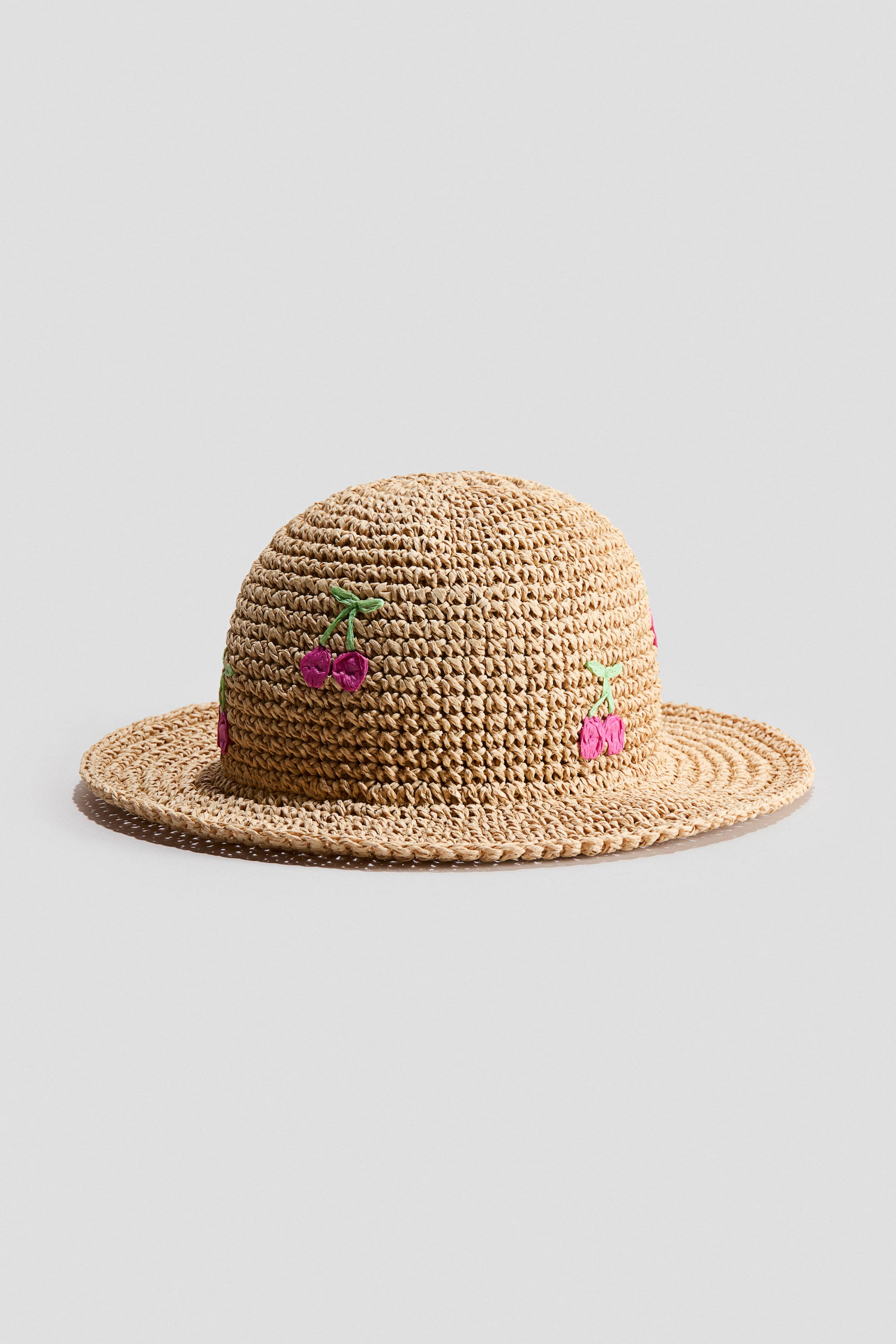 Ampliar la imagen: Un sombrero de paja tejido muestra una corona redondeada y un ala plana, colocado mirando hacia adelante y ligeramente hacia la derecha. Cerezos bordados en rosa con tallos verdes adornan su corona.