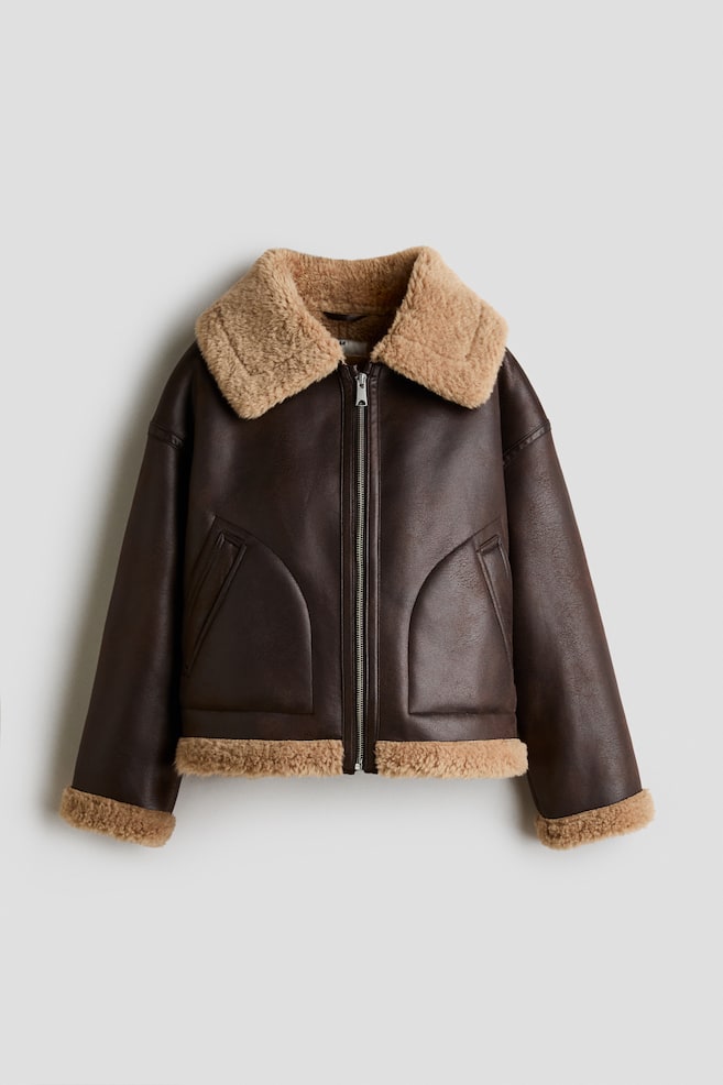 Eine dunkelbraune Aviator-Jacke aus Kunstleder ist von vorn zu sehen und verfÃ¼gt Ã¼ber einen flauschigen, hellbraunen Shearling-Kragen, passende Shearling-BesÃ¤tze an den ÃrmelbÃ¼ndchen und am Saum sowie einen silberfarbenen FrontreiÃverschluss. Die Jacke zeigt eine kastige Silhouette mit zwei schrÃ¤gen Leistentaschen auf der Vorderseite. - 1