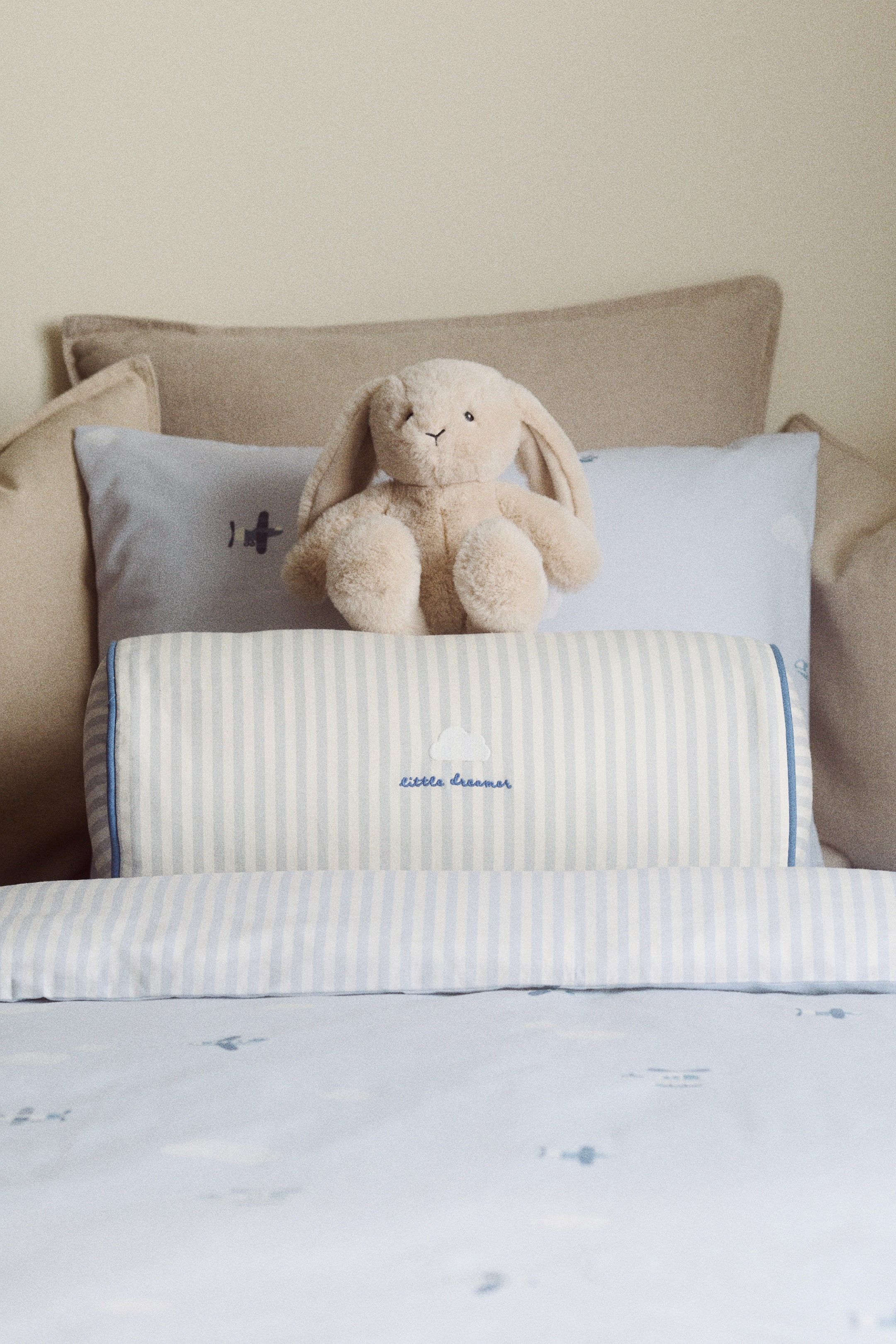 View larger image: A bolster is displayed front-facing, made from cotton canvas and showcasing a striped pattern of light blue and white vertical lines. It features a blue embroidered detail depicting a cloud shape above the text 'little dreamer', and is finished with blue piping along its sides.